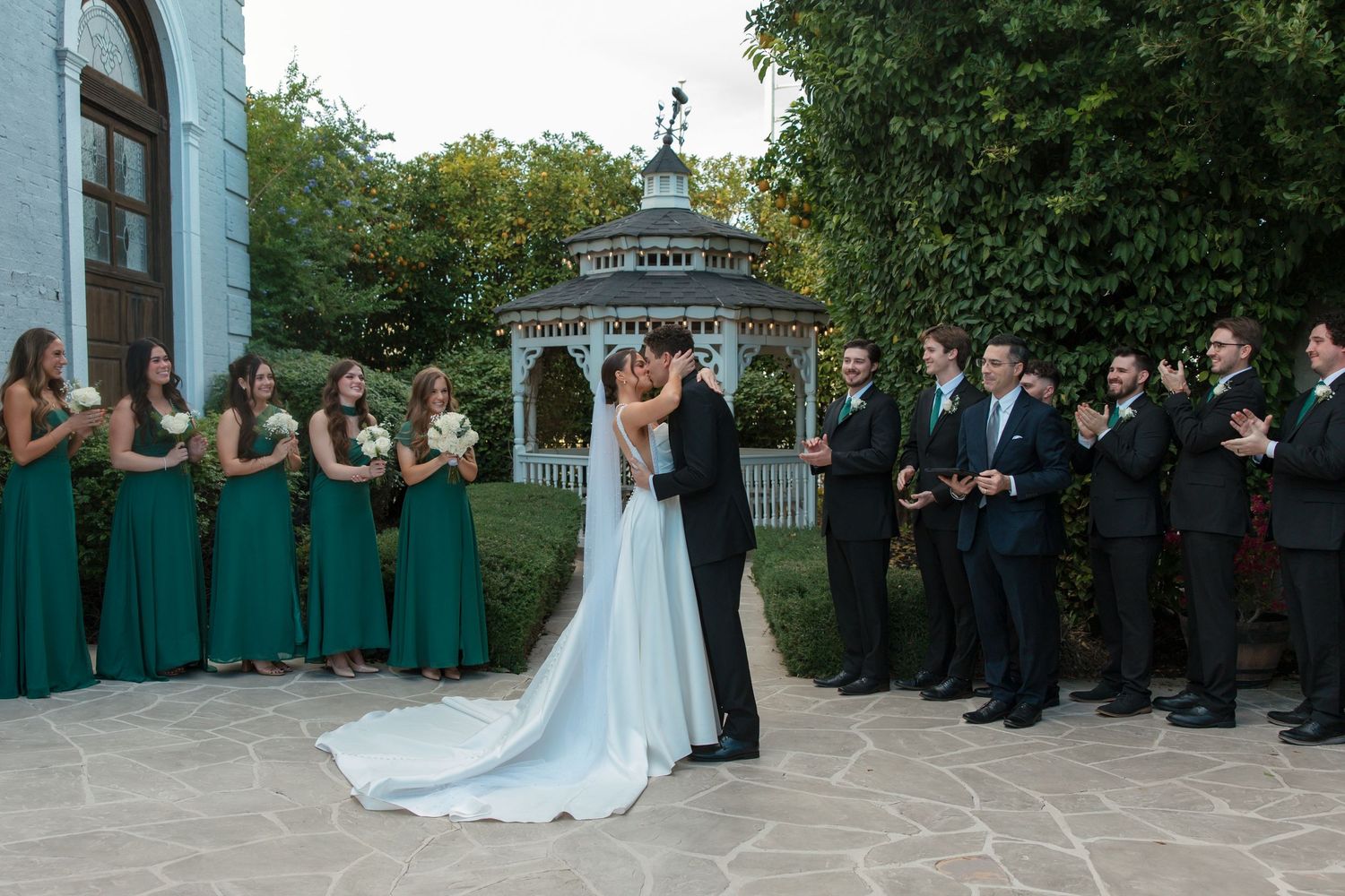 Newlyweds share first kiss surrounded by wedding party in emerald green dresses near gazebo.