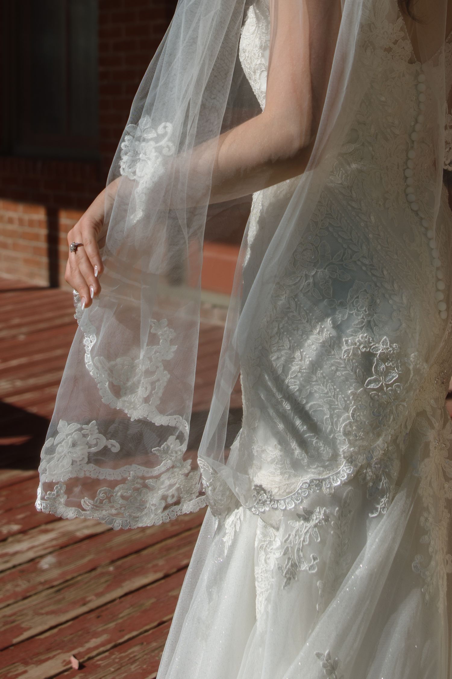 Close-up of a brides lace wedding dress and veil detail in warm sunlight on a wooden deck.