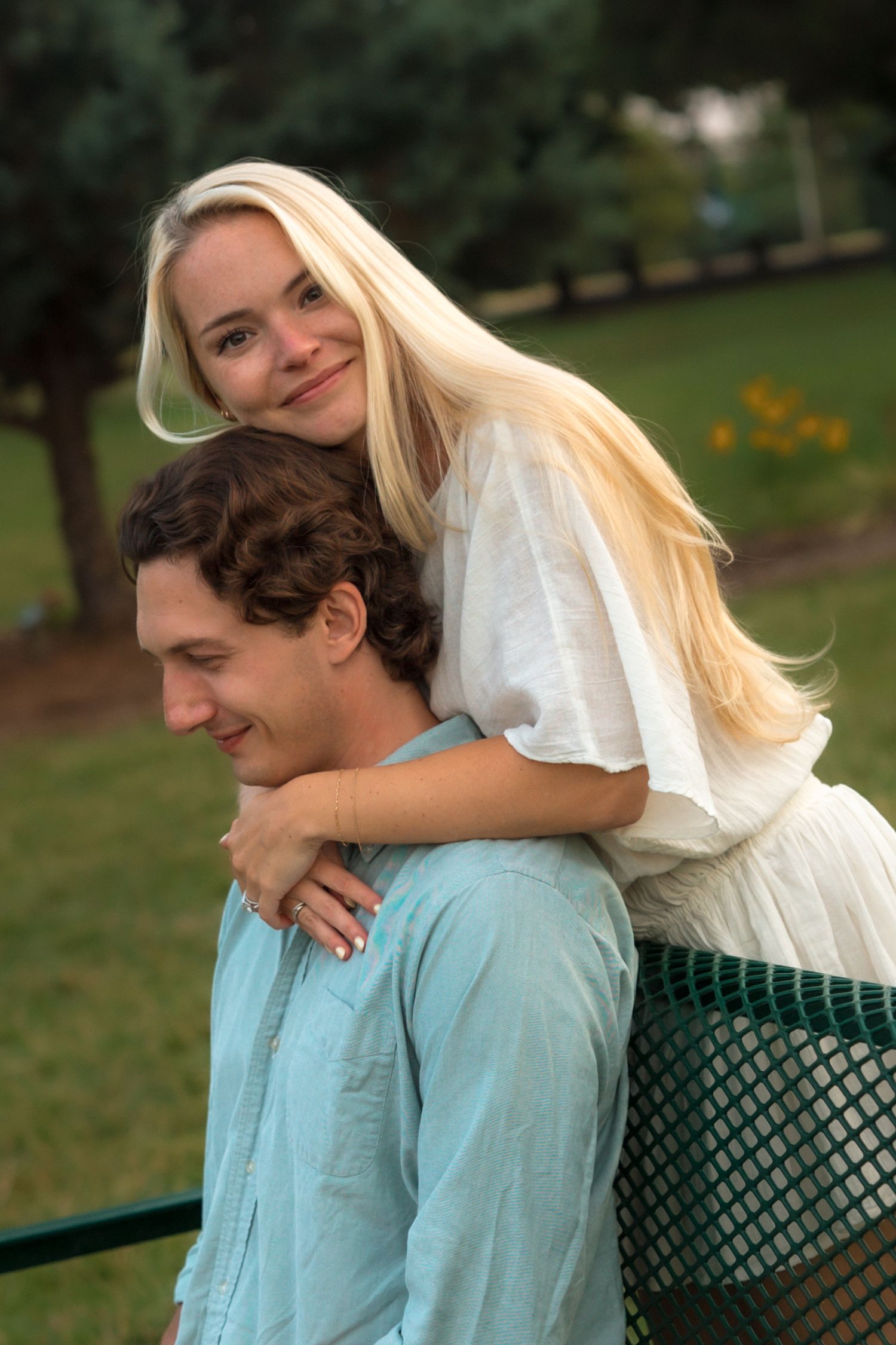 Smiling blonde woman leaning over man's shoulders from behind a green bench in a sunny park.