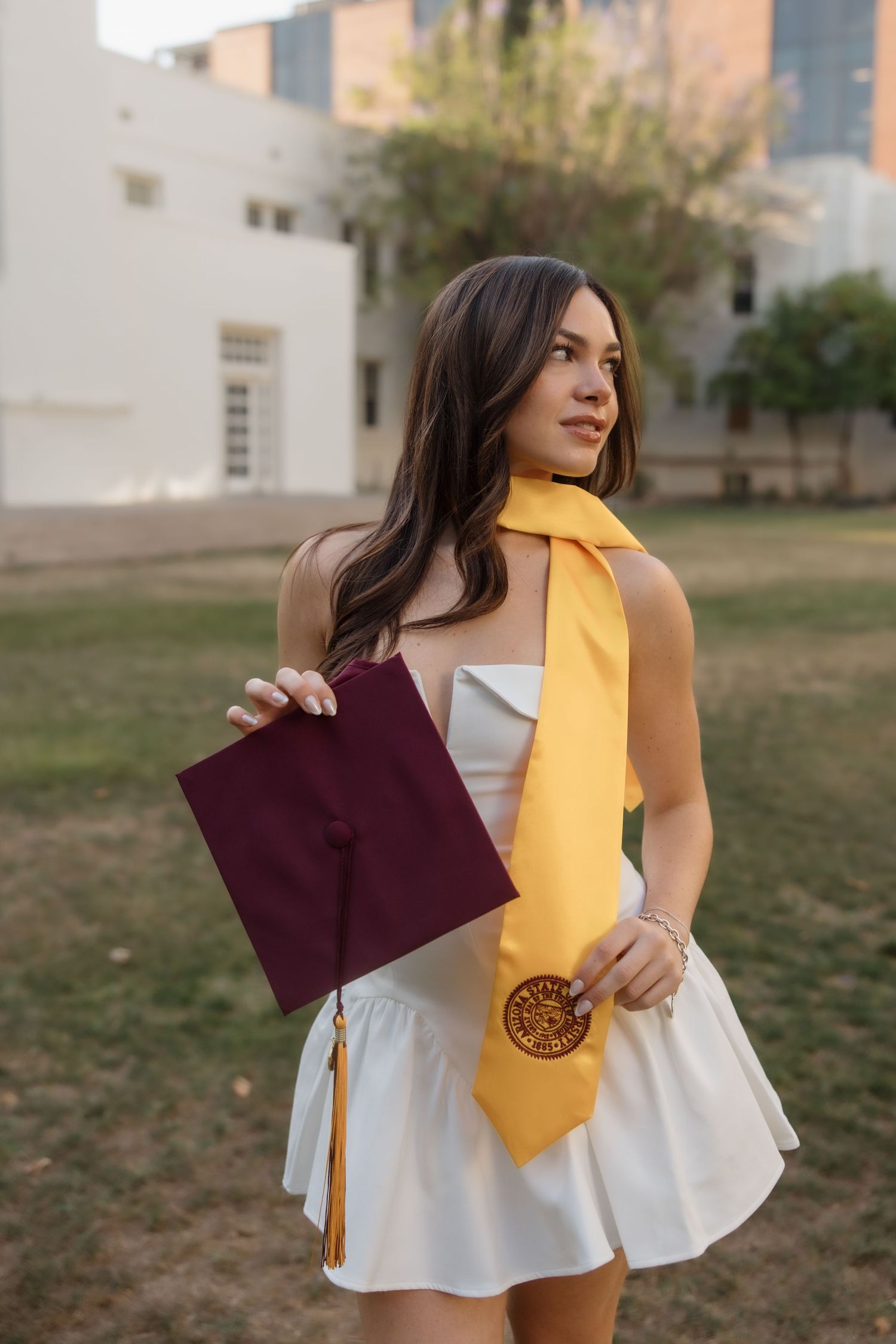 Young woman in white dress holding maroon graduation cap with gold stole outdoors on campus grounds.