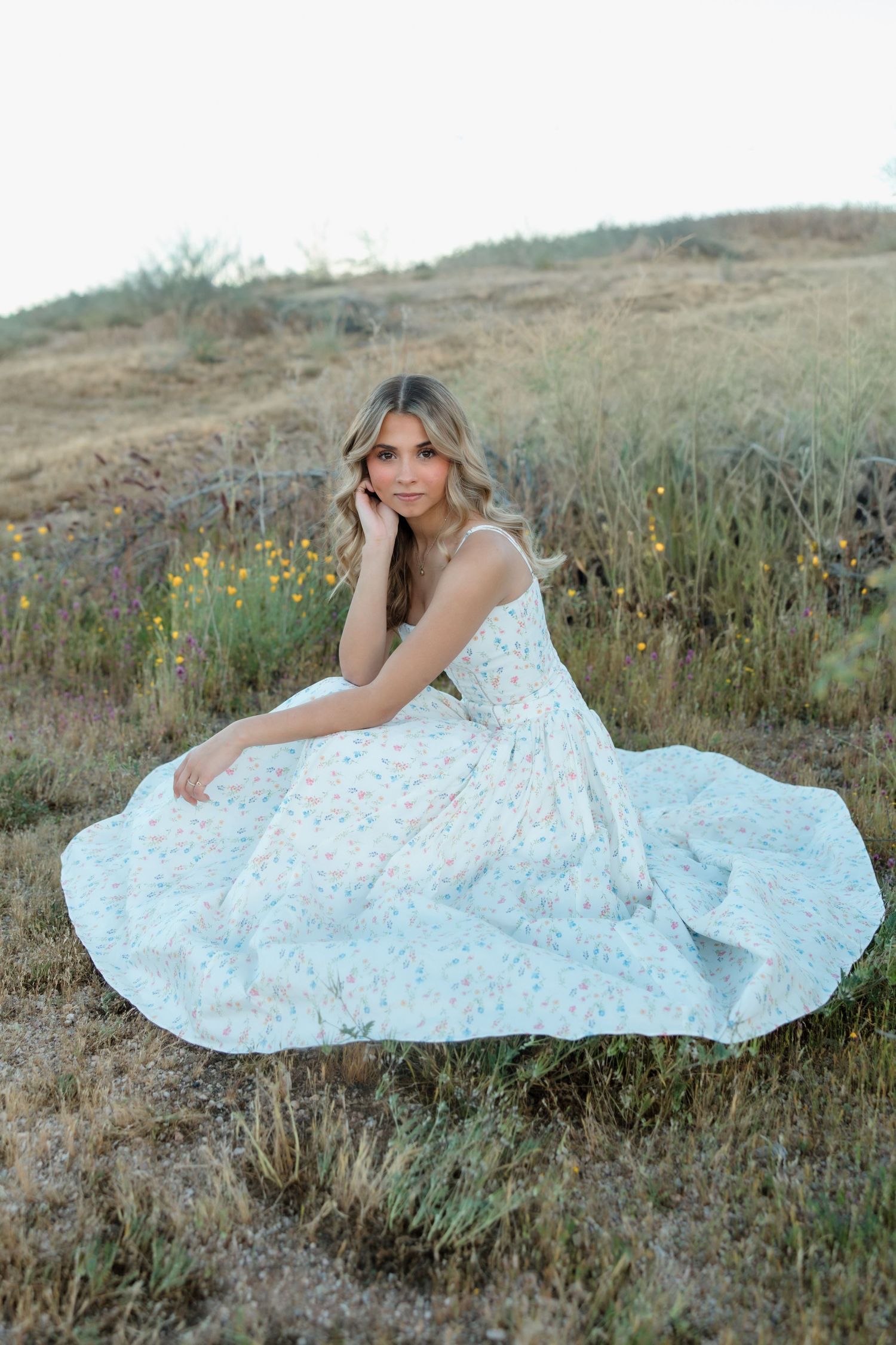 Beautiful young woman in white floral maxi dress sitting gracefully in wildflower field at golden hour.