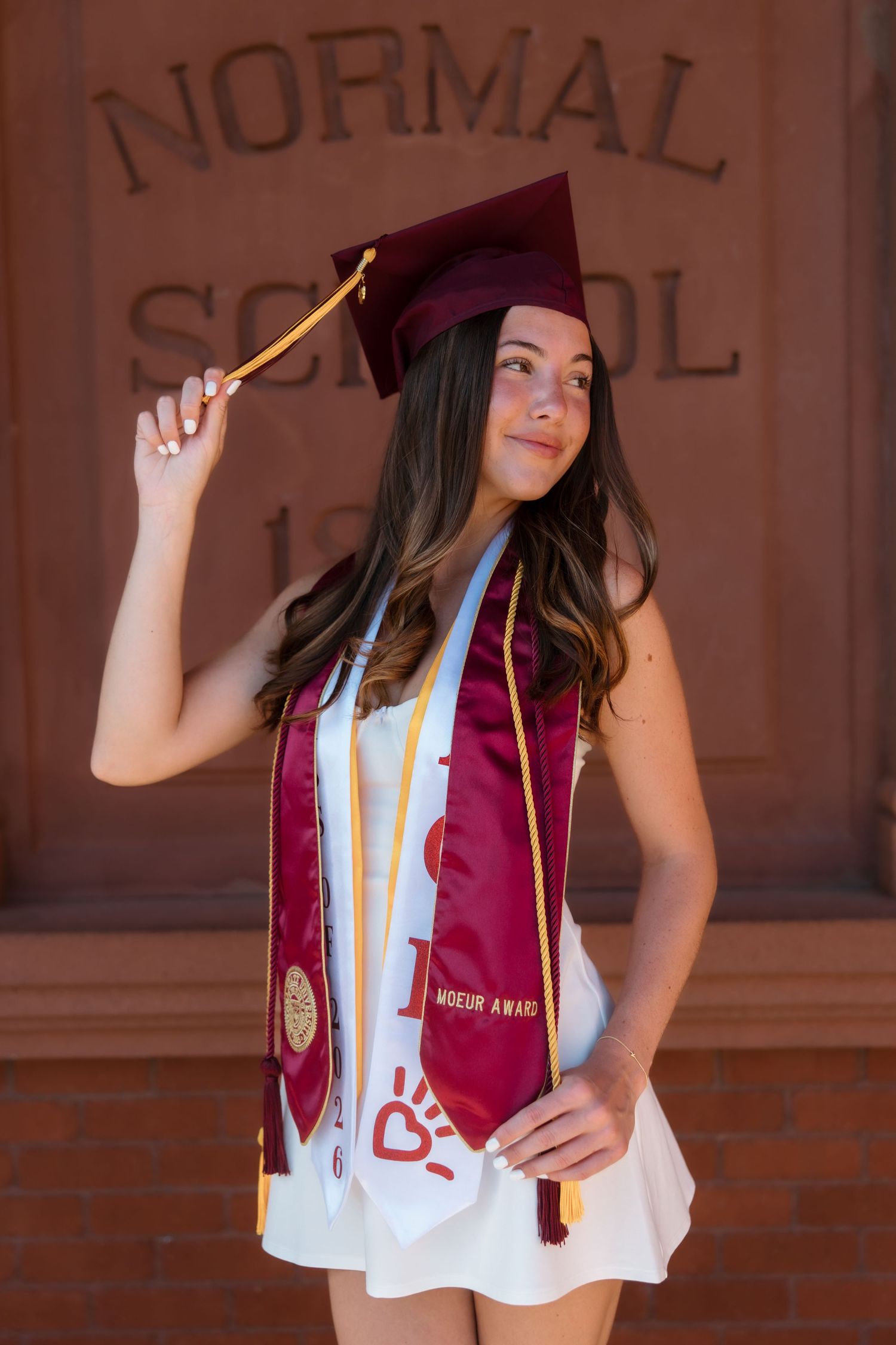 Proud female graduate in maroon cap and honor stoles posing before historic Normal School building.