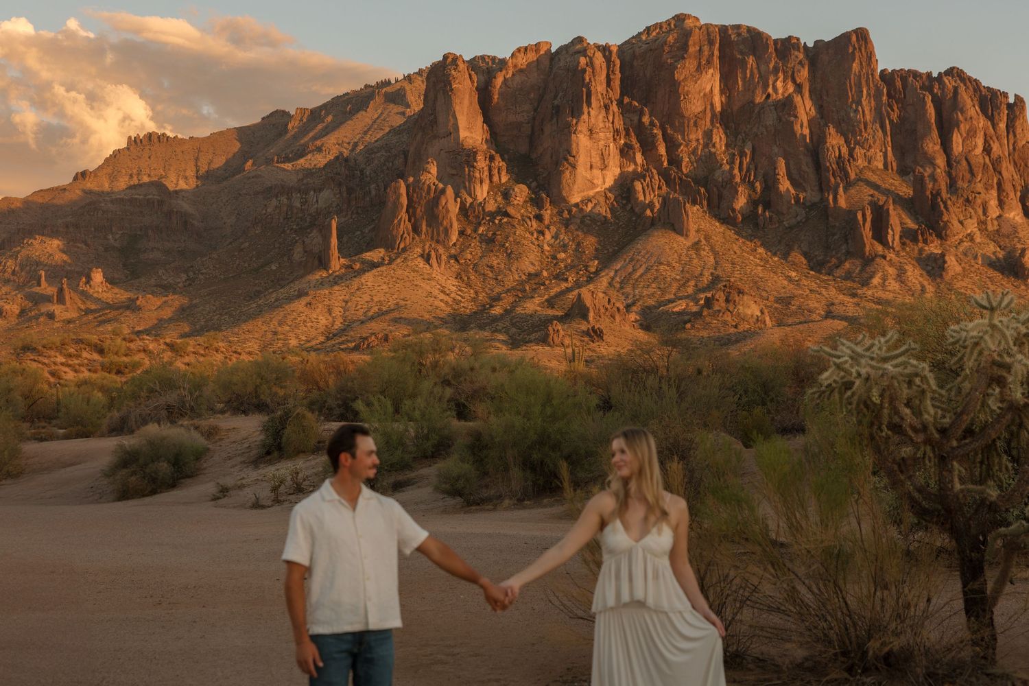 Couple holding hands at sunset in Arizona desert with Superstition Mountains glowing orange in background.