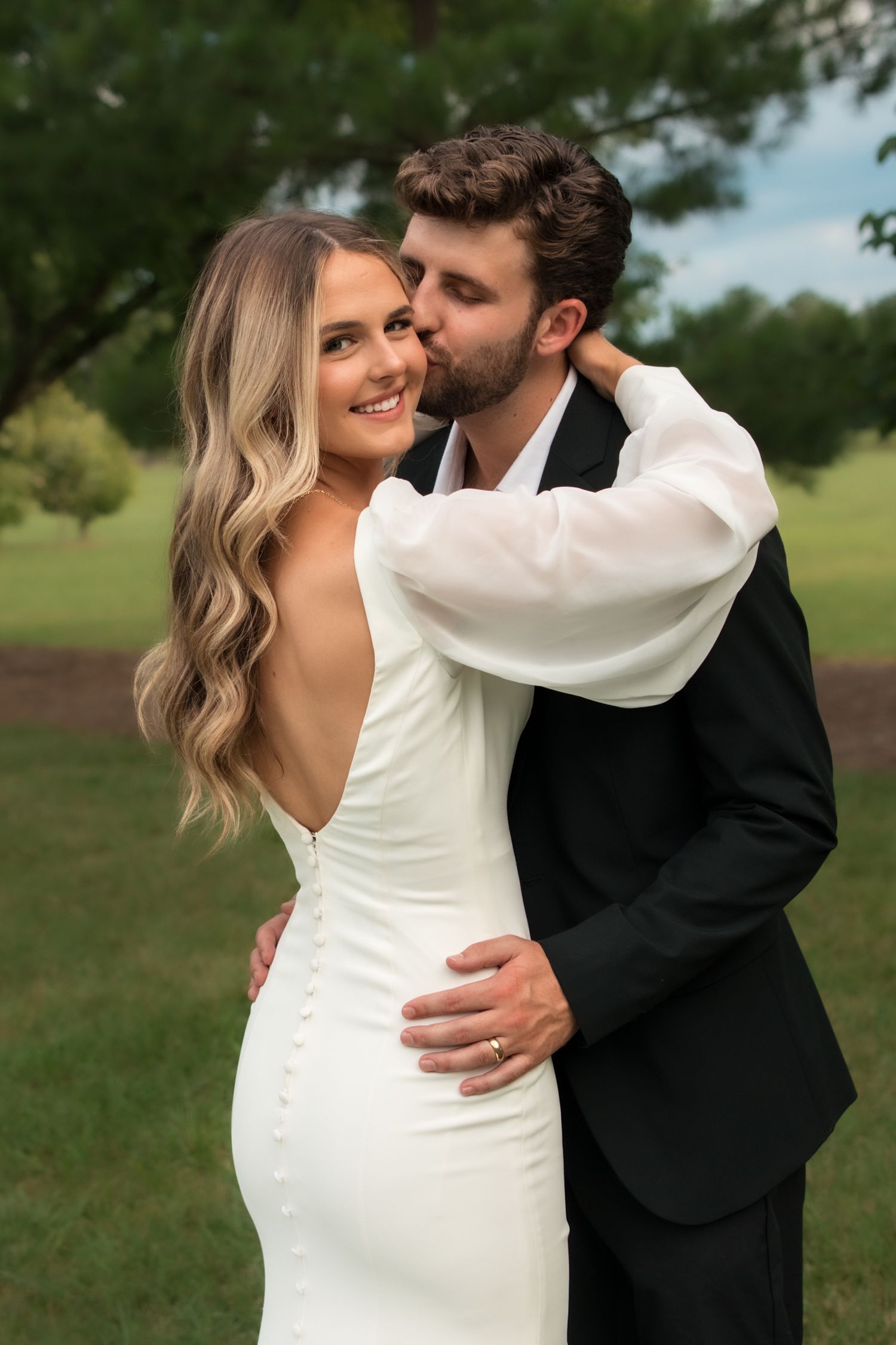 Groom kissing smiling bride on cheek outdoors, bride wearing backless white fitted gown with button details.