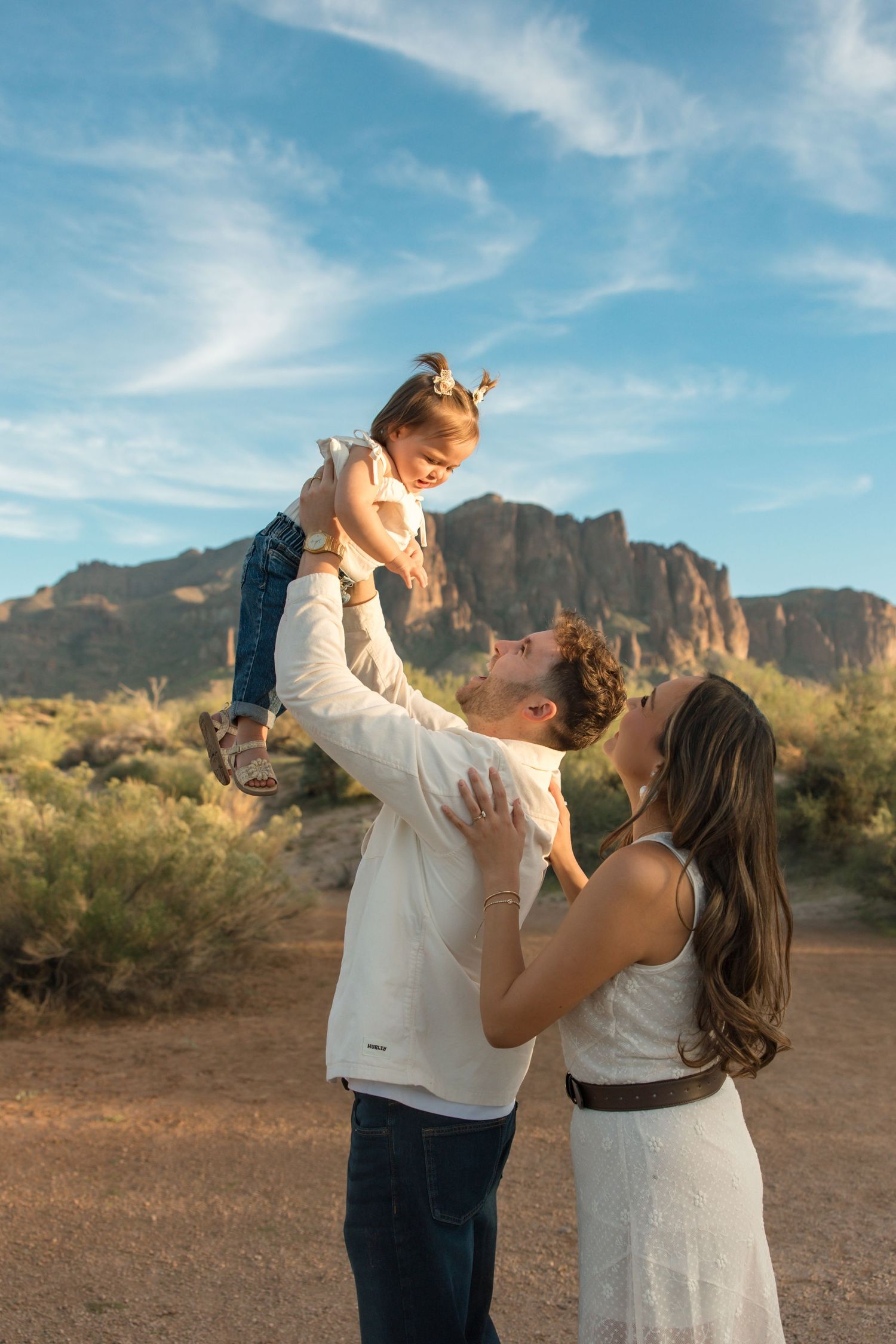A couple lifts their toddler in the Arizona desert with Superstition Mountains backdrop on a sunny day.
