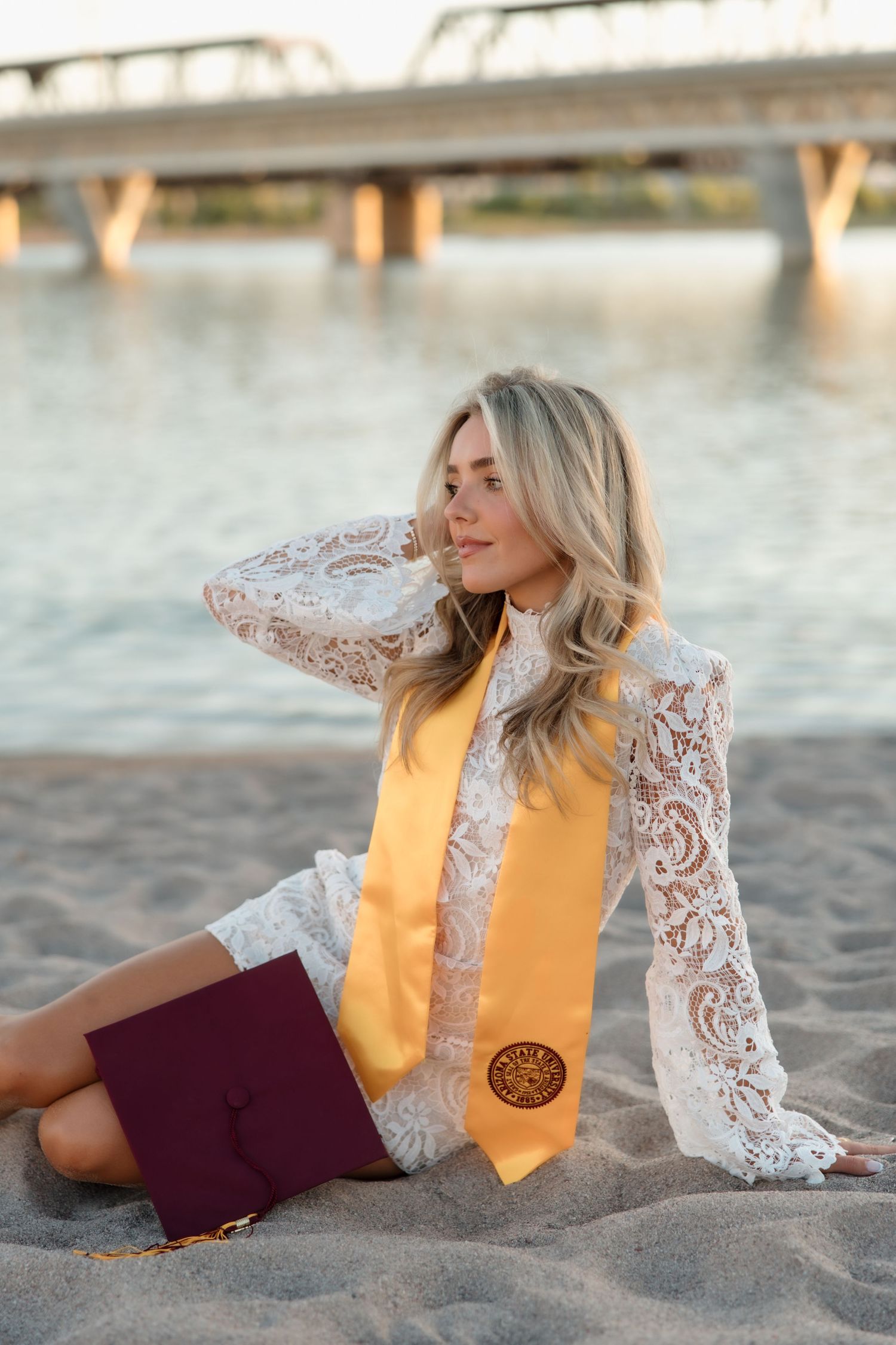 Graduate in white lace dress and gold stole sitting on sandy beach holding maroon cap at sunset.