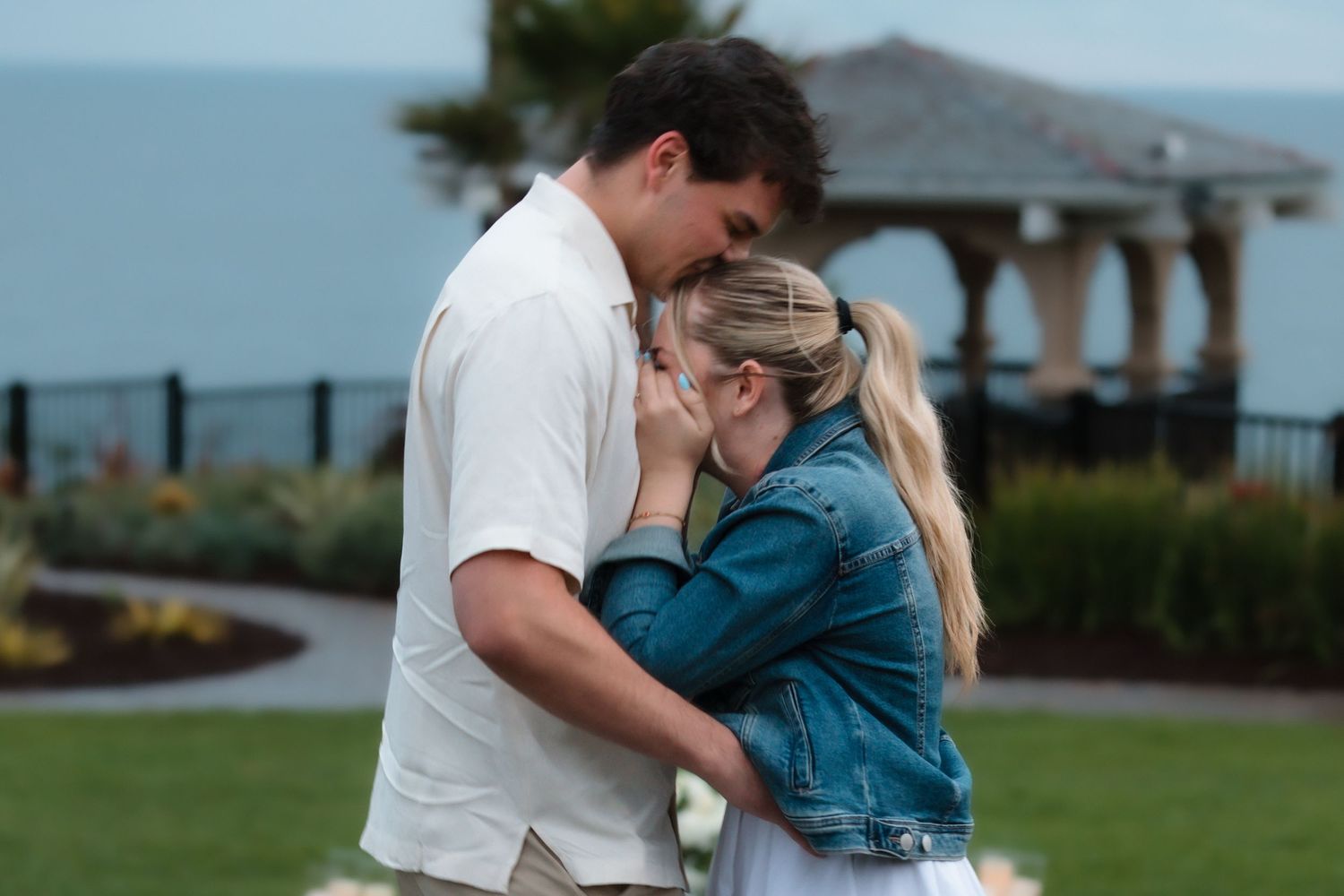 Couple embracing romantically outdoors near coastal gazebo, man in white shirt hugging emotional blonde woman in denim jacket.