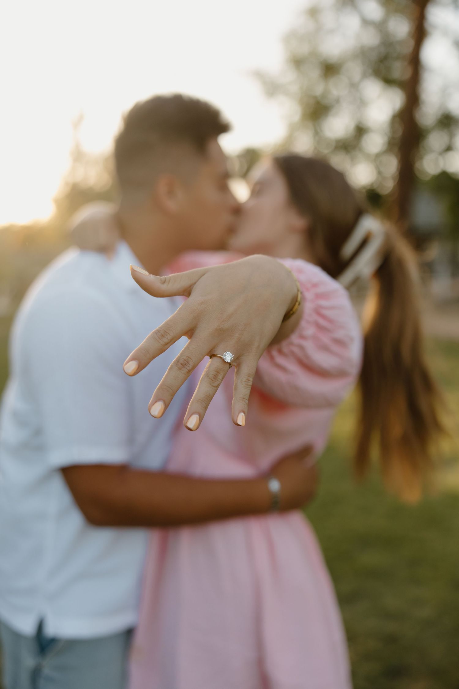 Couple kissing at sunset while showing off engagement ring, woman in pink dress embracing man in white shirt outdoors.