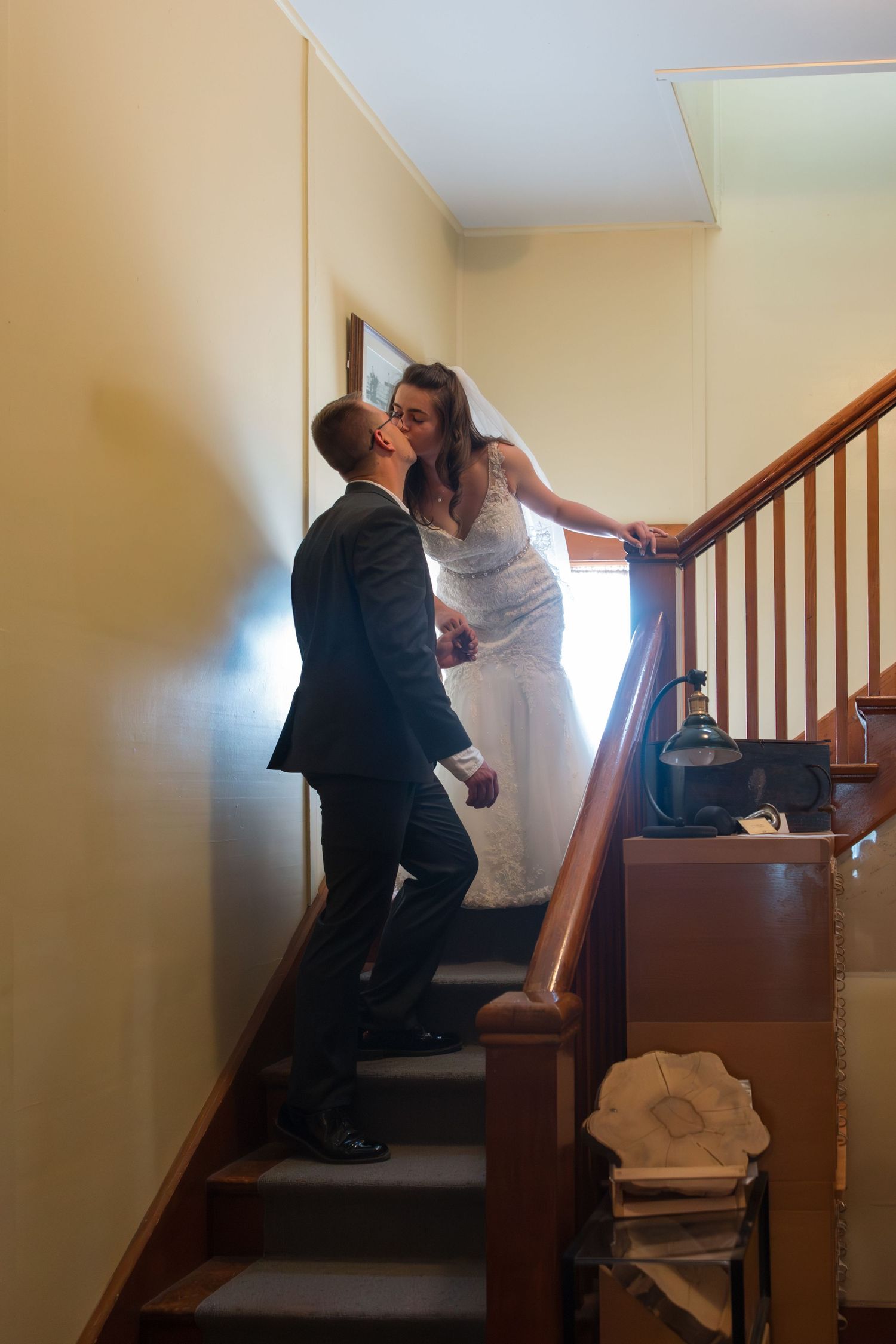 Bride in white gown and groom in black suit share a romantic moment on a wooden staircase indoors.