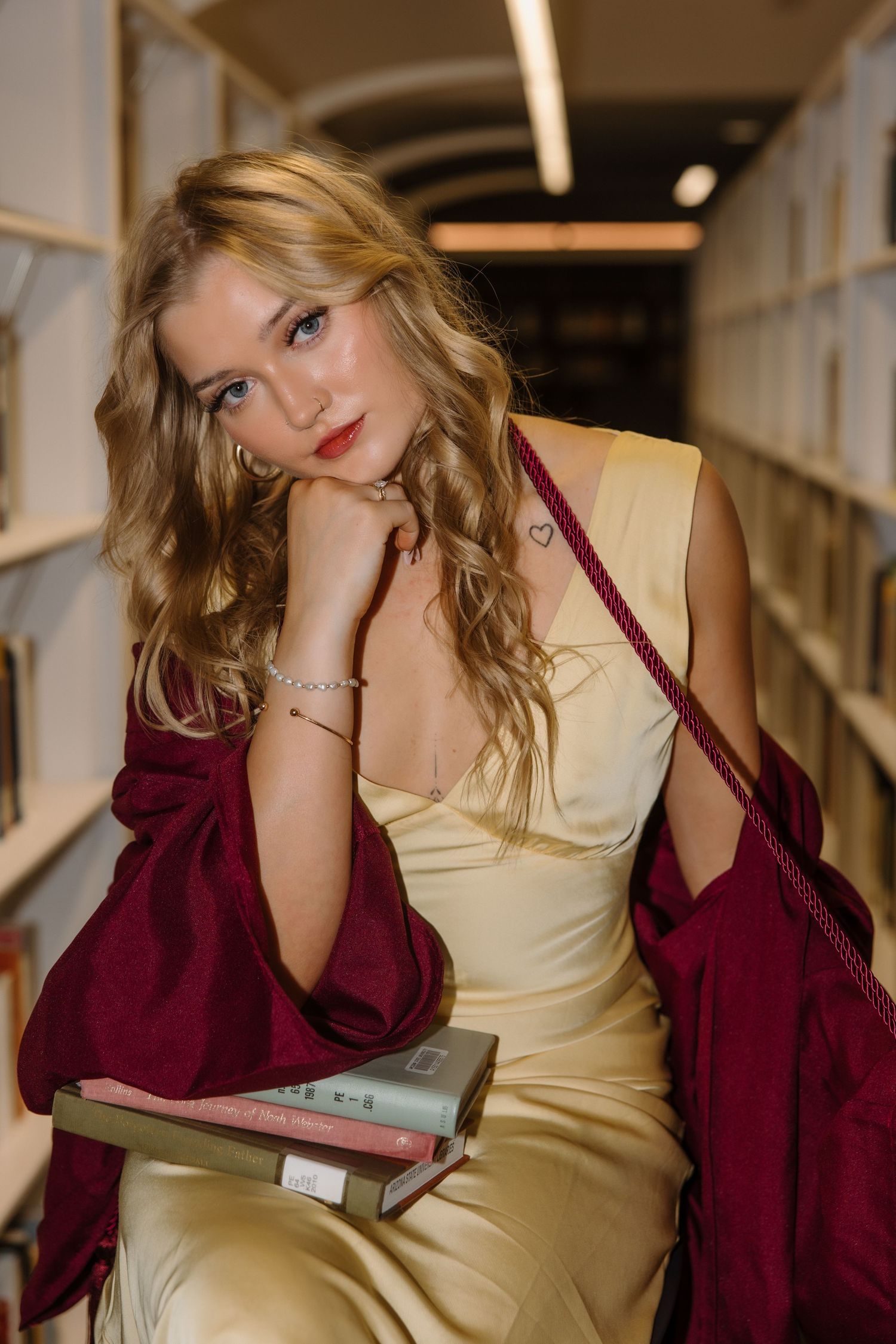 Blonde woman in gold dress holding books in library aisle with burgundy wrap.