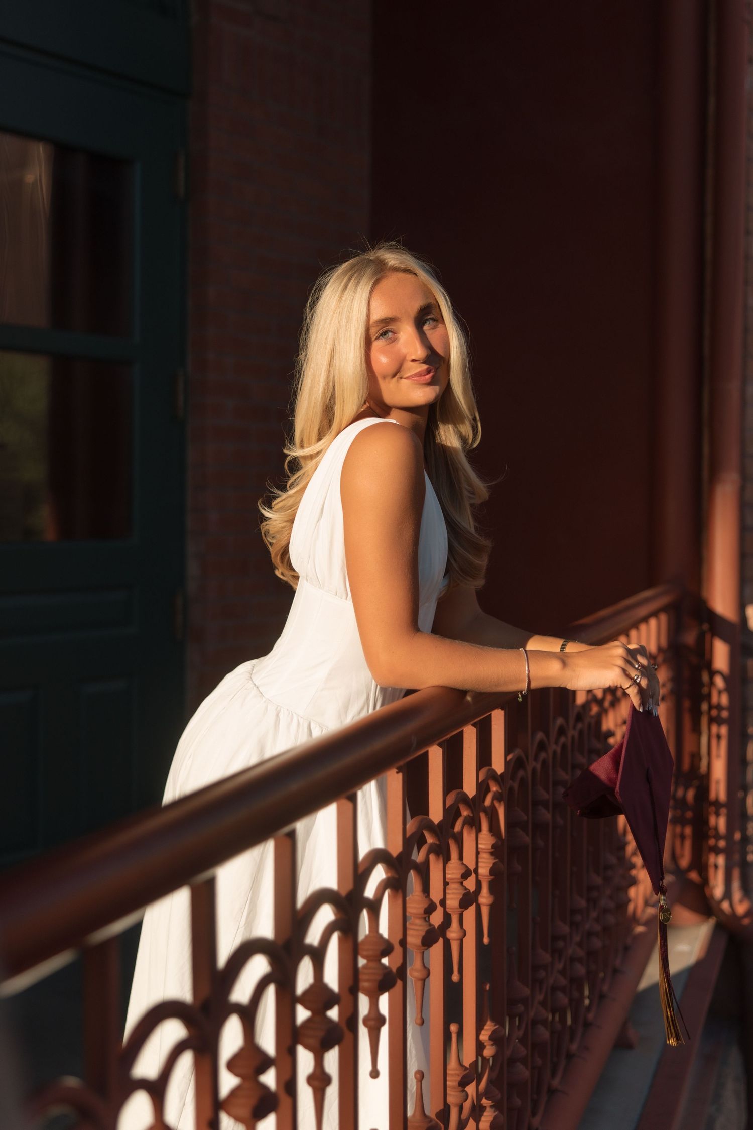 Blonde woman in white sleeveless dress leaning on ornate railing in warm golden sunset light.