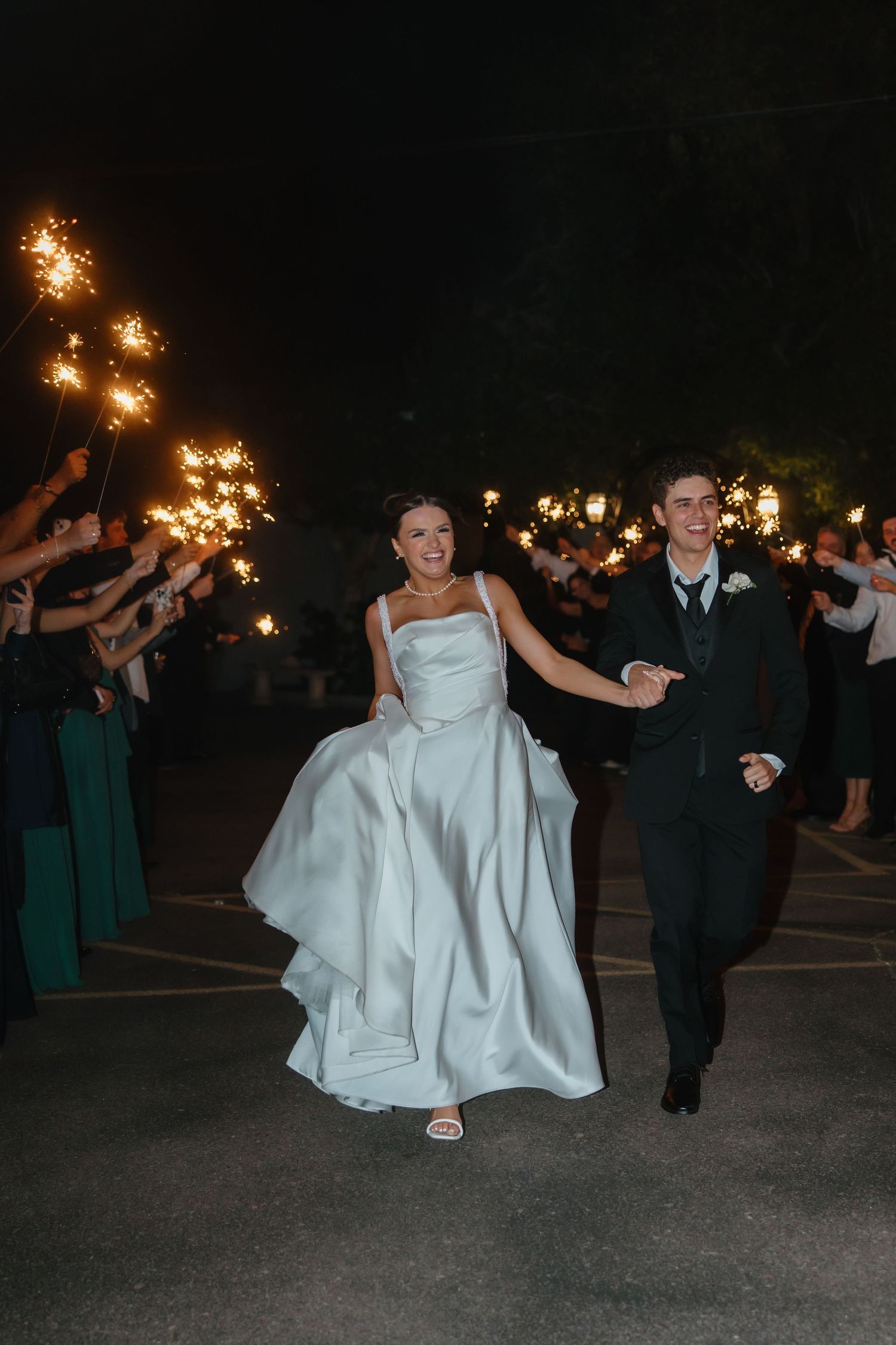 Bride and groom run joyfully through sparkler exit tunnel held by wedding guests at night celebration.