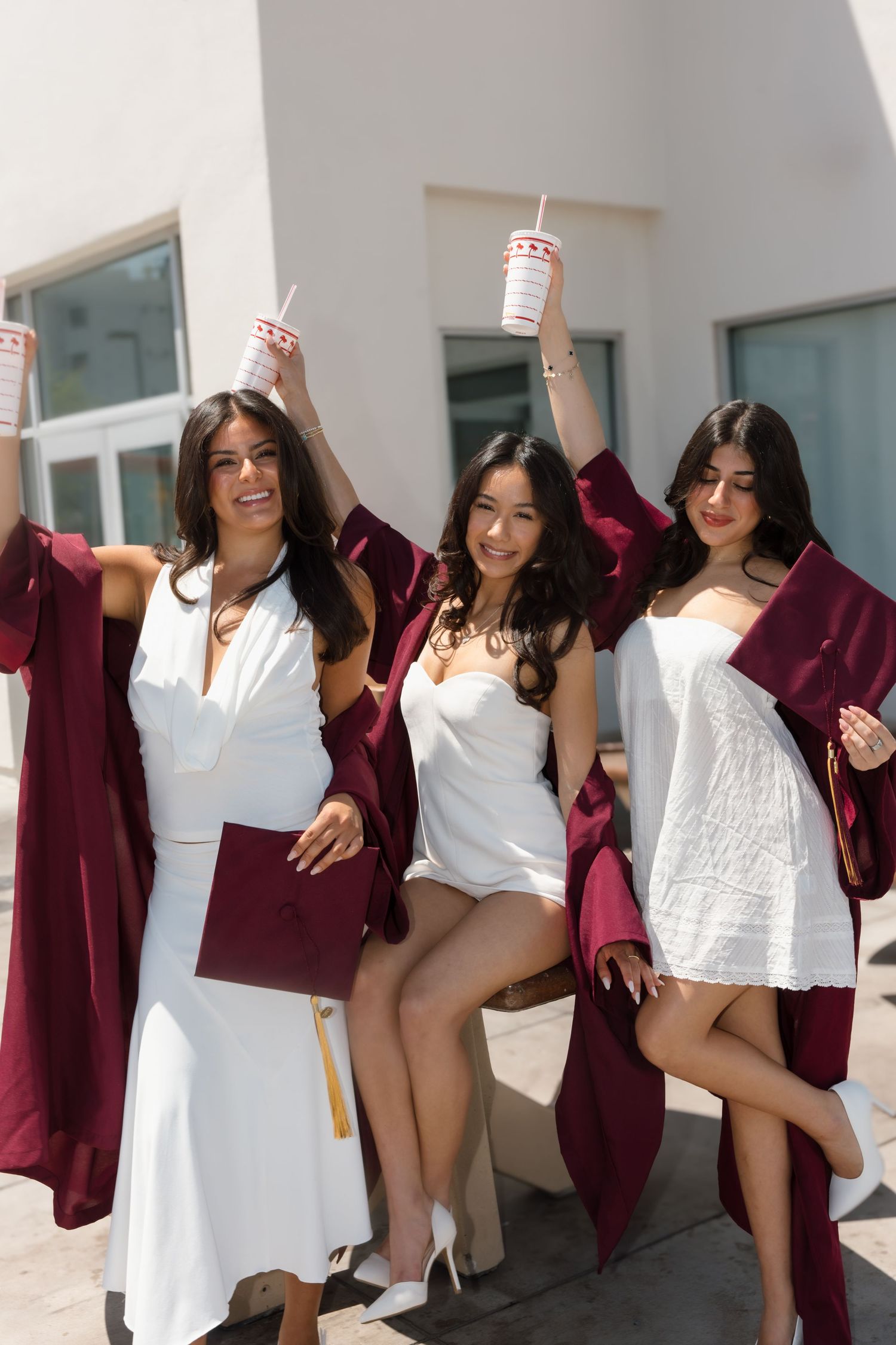 Three smiling female graduates in burgundy robes raise In-N-Out drinks while sitting outside the restaurant.