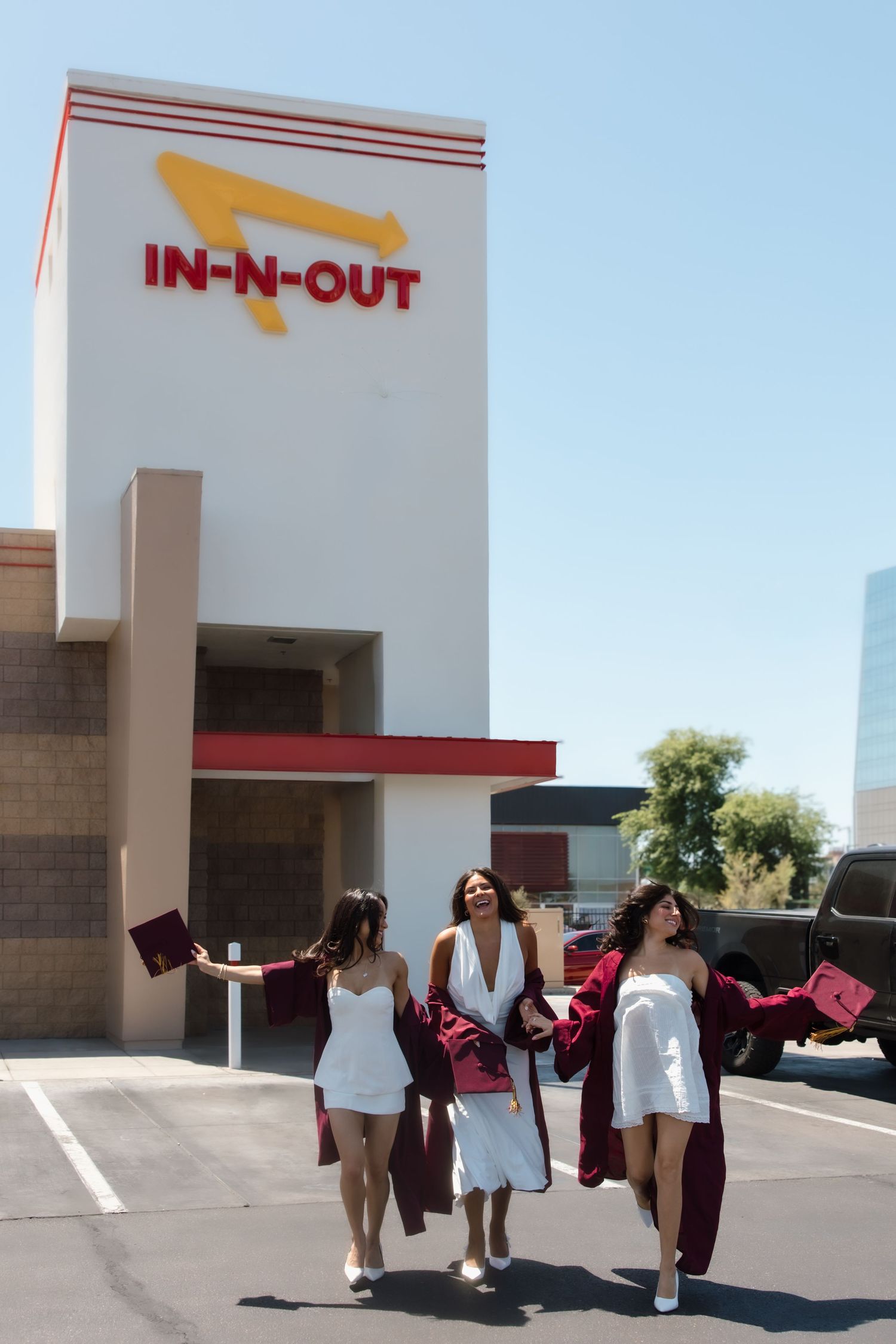 Three graduates in white dresses and maroon gowns walking in front of In-N-Out Burger.