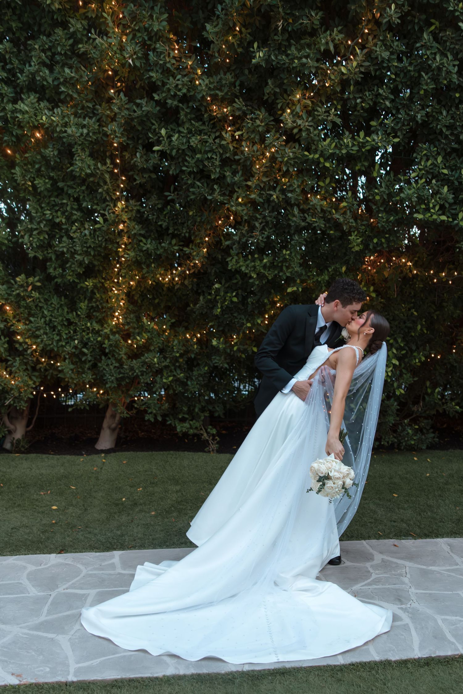 Groom dips bride for a romantic kiss in front of a beautifully lit hedge at dusk.