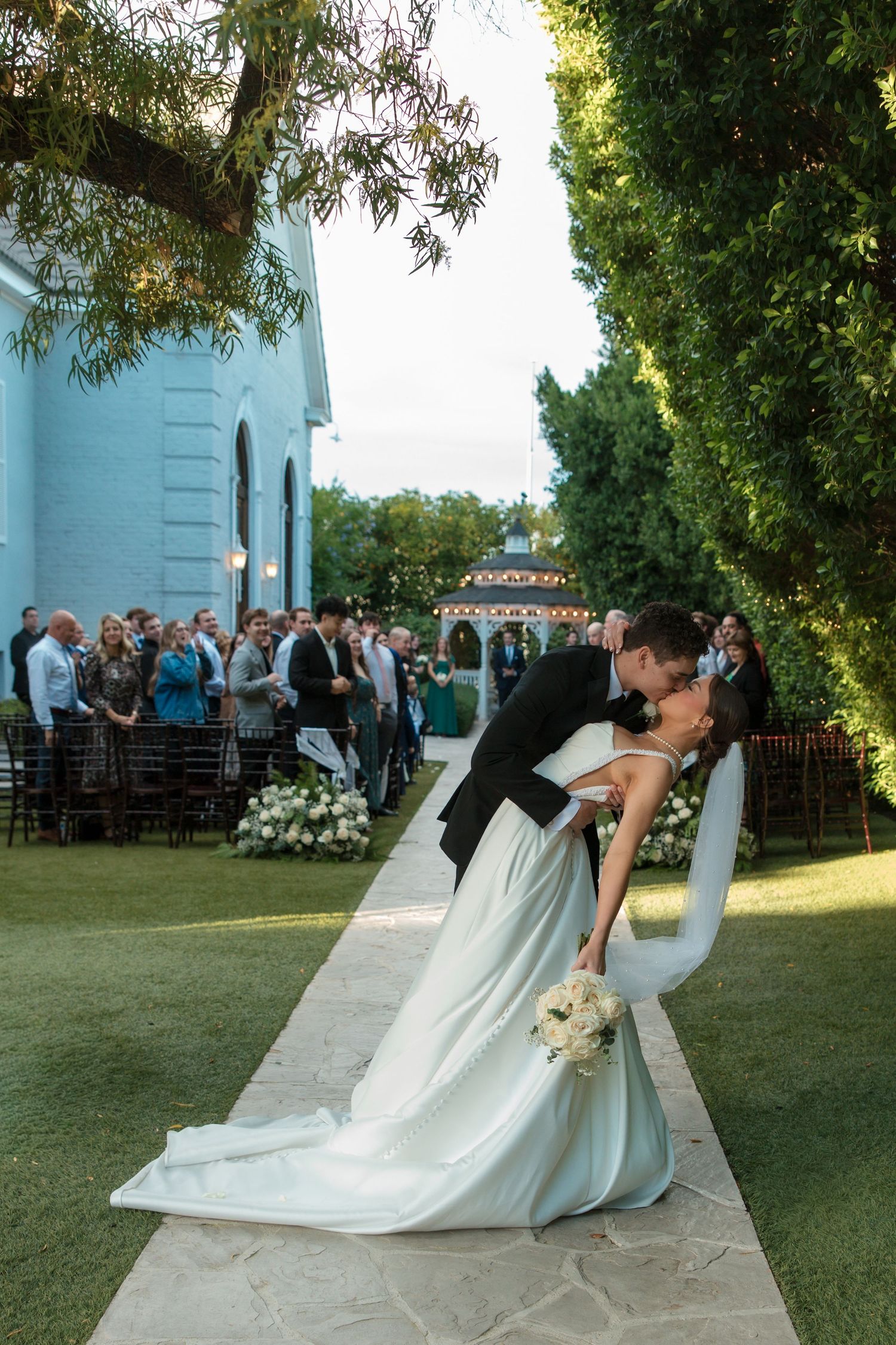 Groom dips bride for romantic kiss on garden pathway with wedding guests applauding behind them.