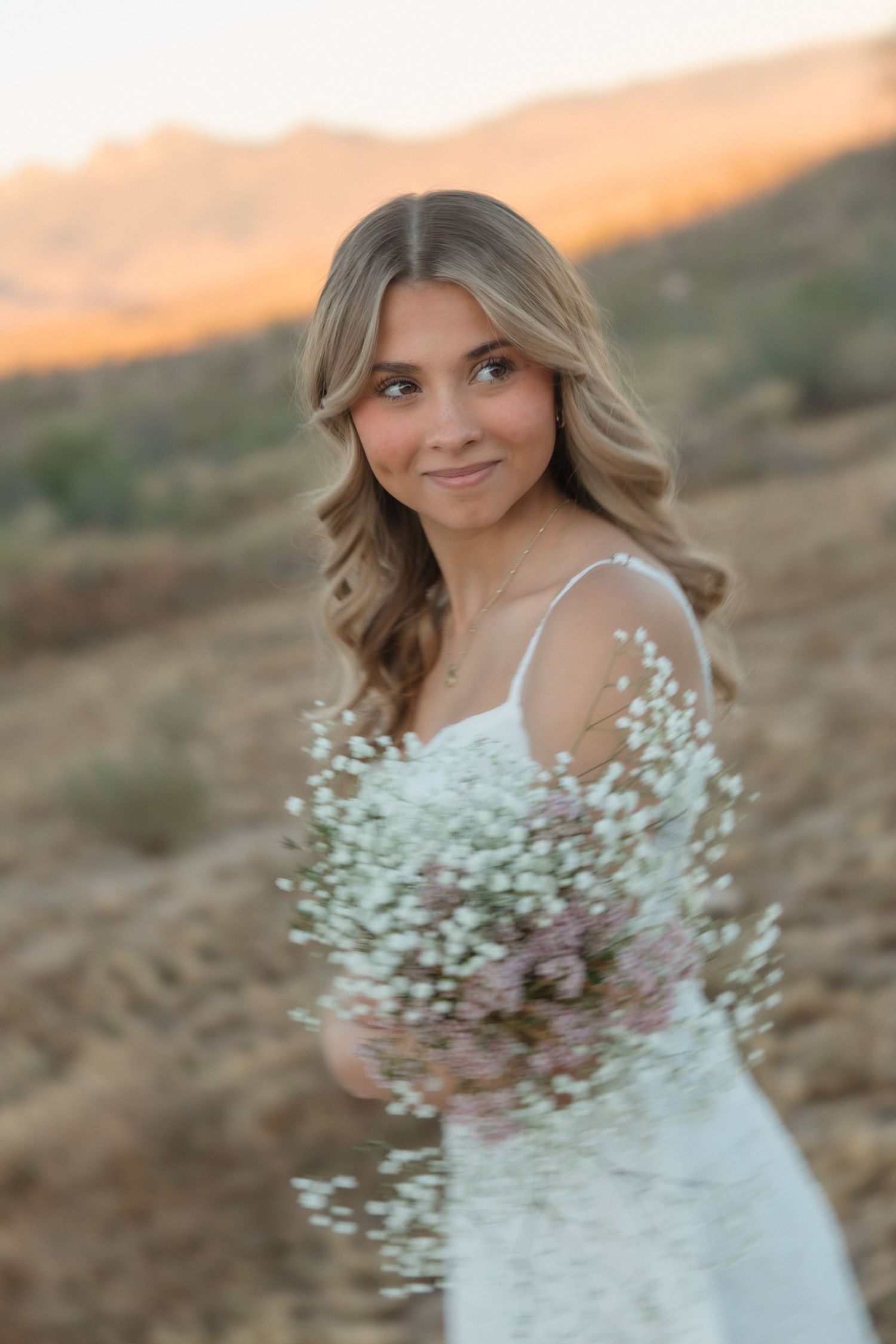 Young woman in white dress holding bouquet of baby's breath flowers at sunset for senior portraits.