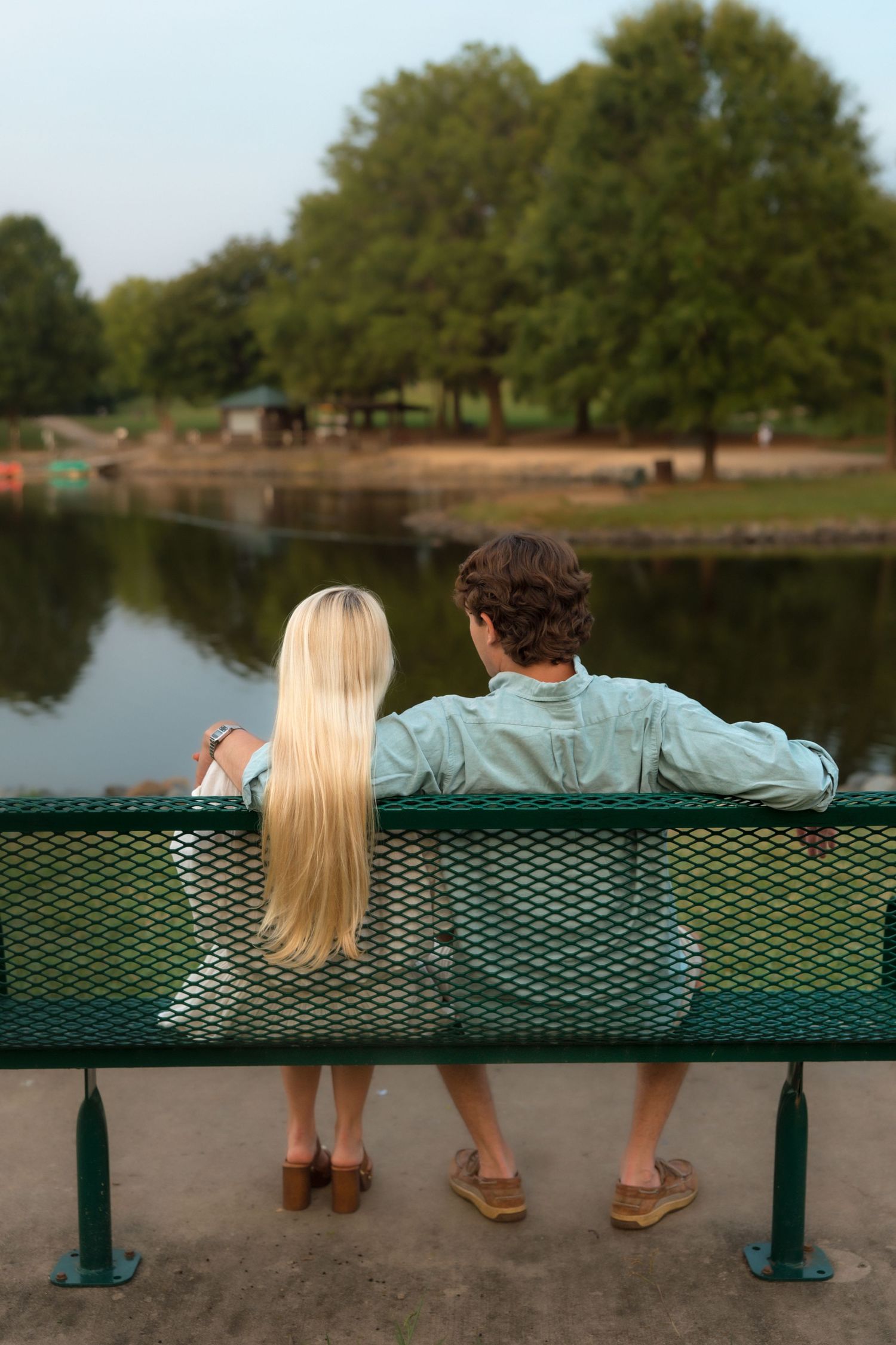 Couple sitting on green park bench from behind, overlooking a serene lake surrounded by lush trees.