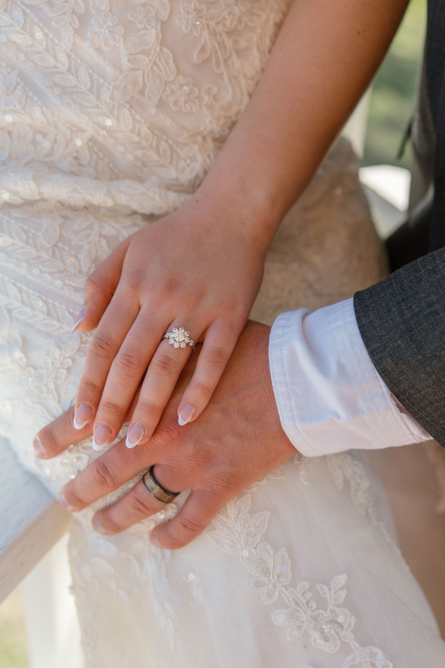 Bride and groom holding hands showing diamond engagement ring and wedding band on lace wedding dress background.