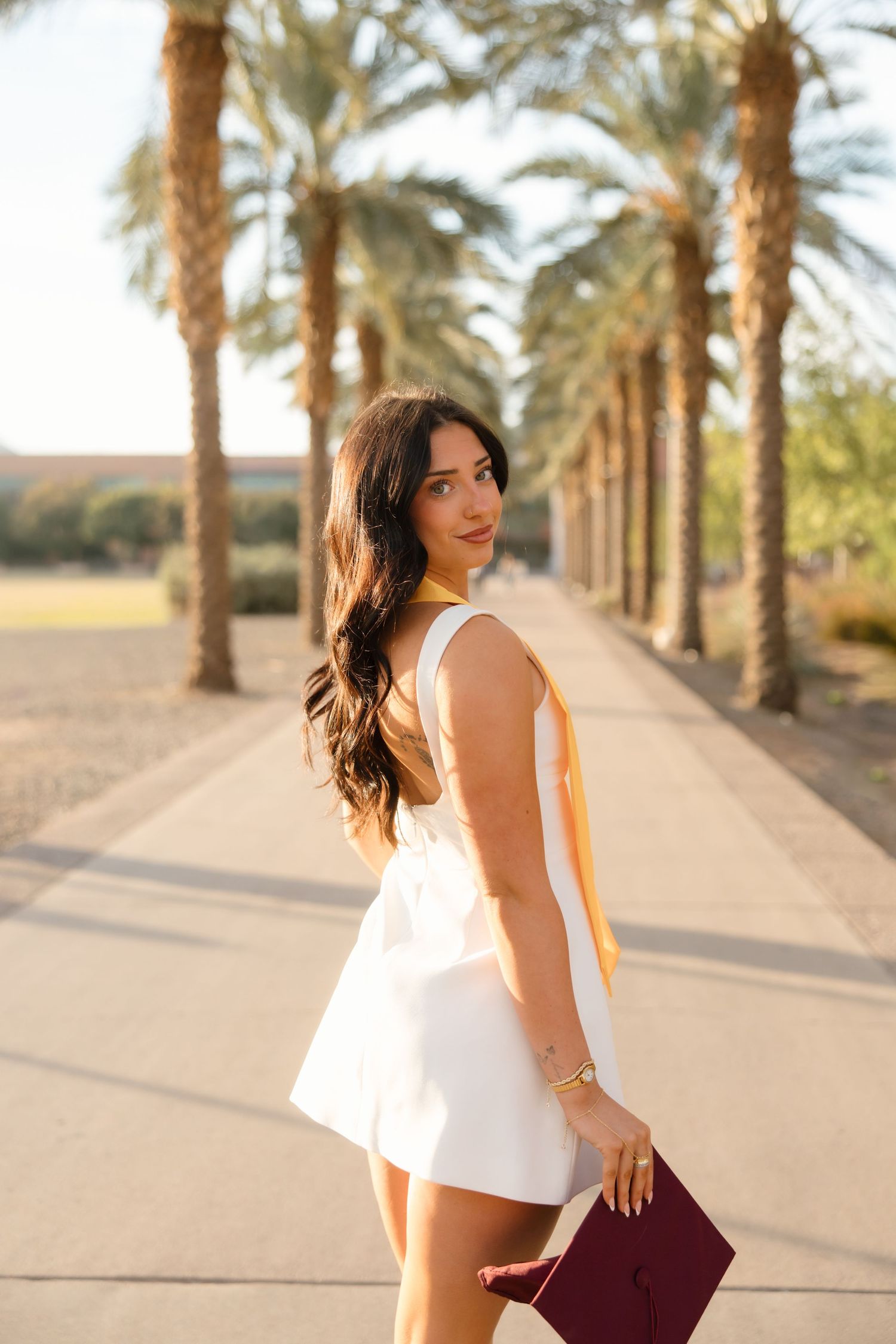 Young woman in white romper holding a maroon graduation cap poses on a palm tree-lined pathway in golden hour light.