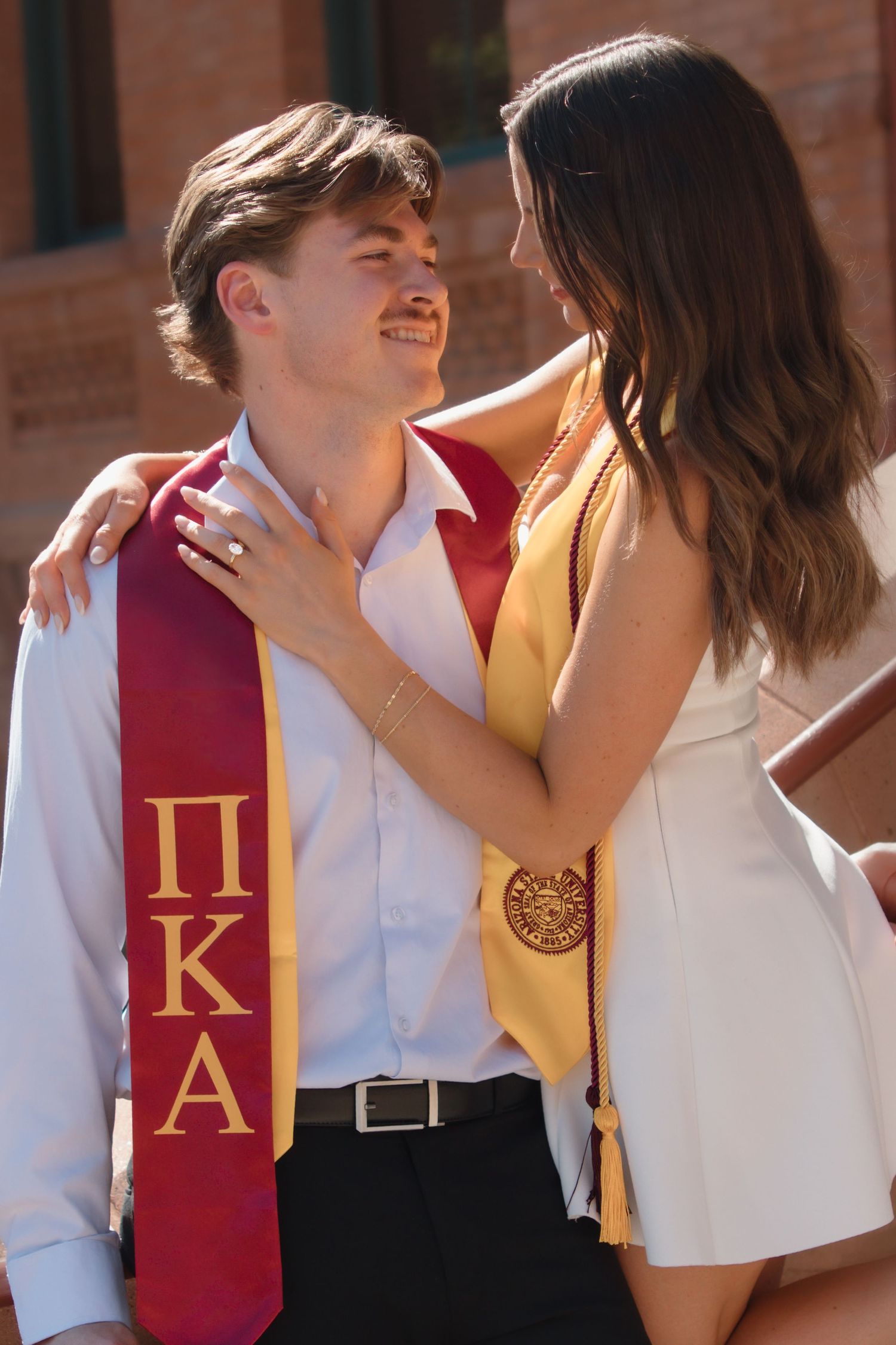 Graduating couple in Pi Kappa Alpha and gold stoles smiling at each other on sunny campus steps.