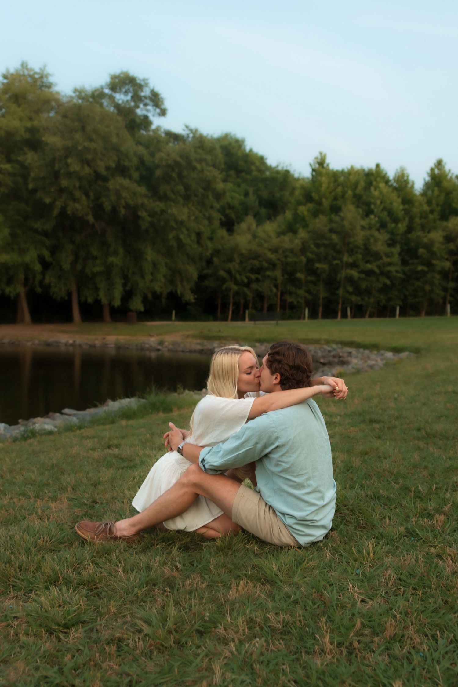 Romantic couple kissing while sitting on grass near a peaceful lake in a park.