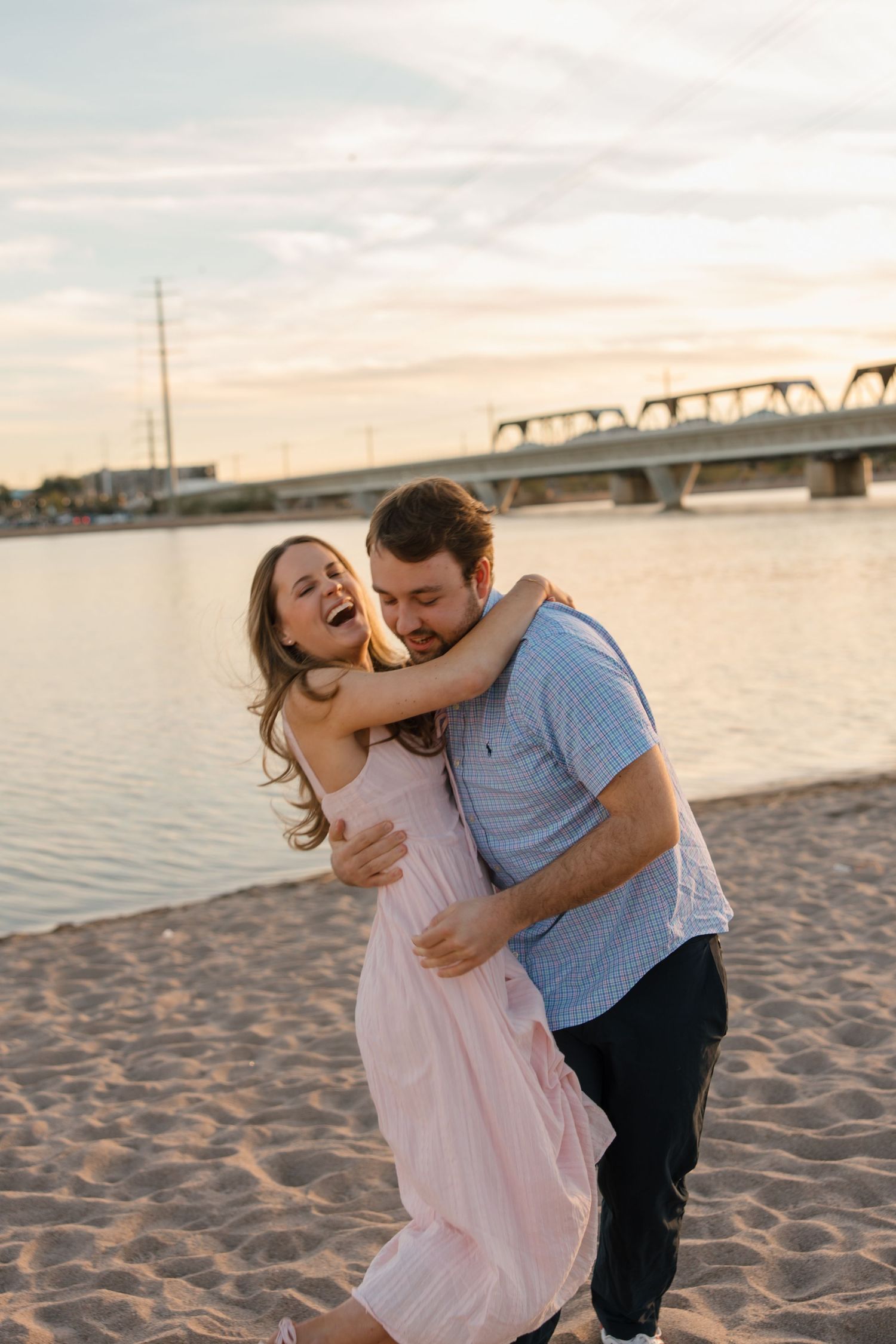 A joyful couple embraces and laughs together on a sandy beach at sunset with a bridge and river in the background.