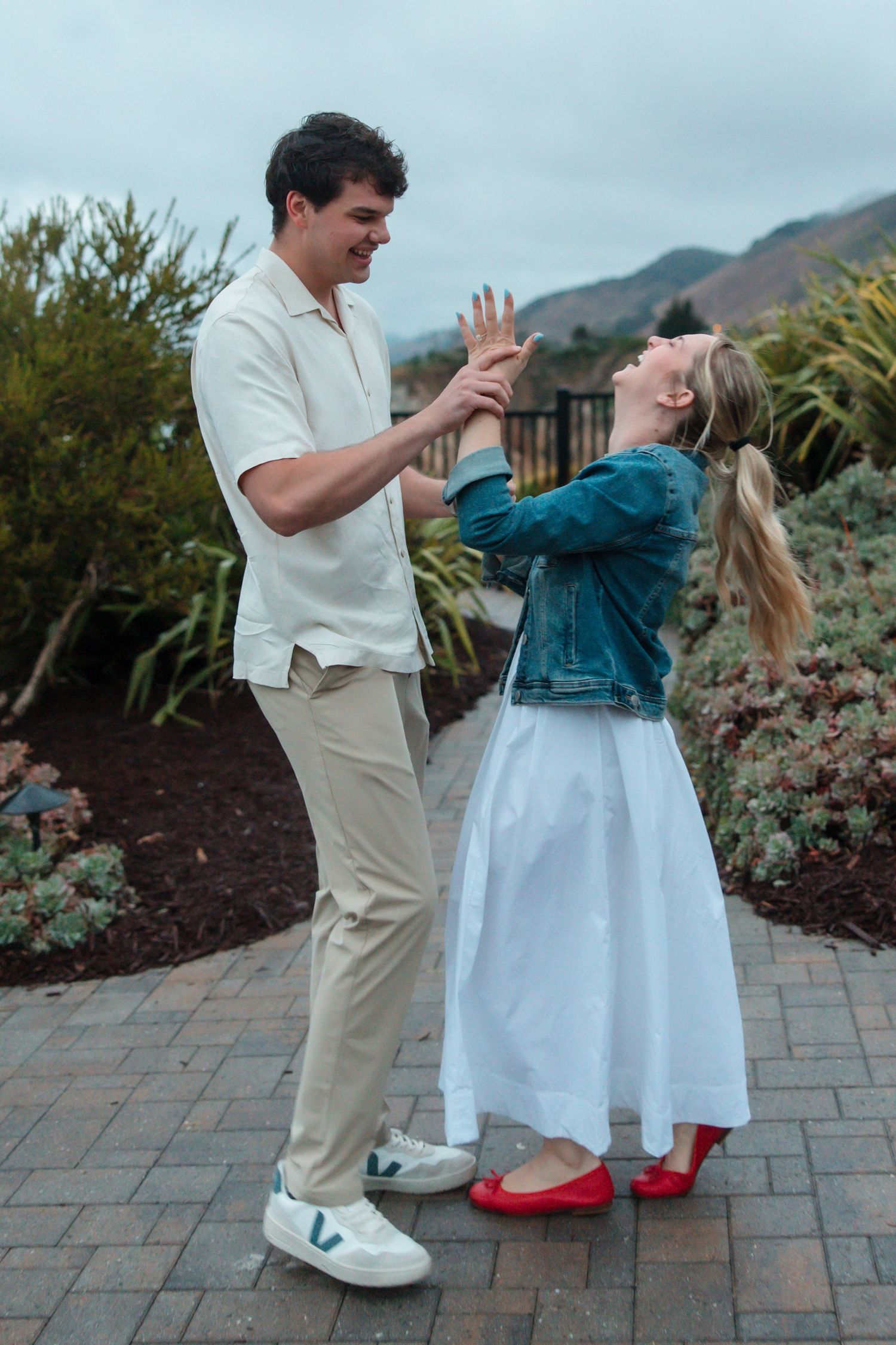 Excited woman admires her new engagement ring while holding hands with her fiance on garden path.