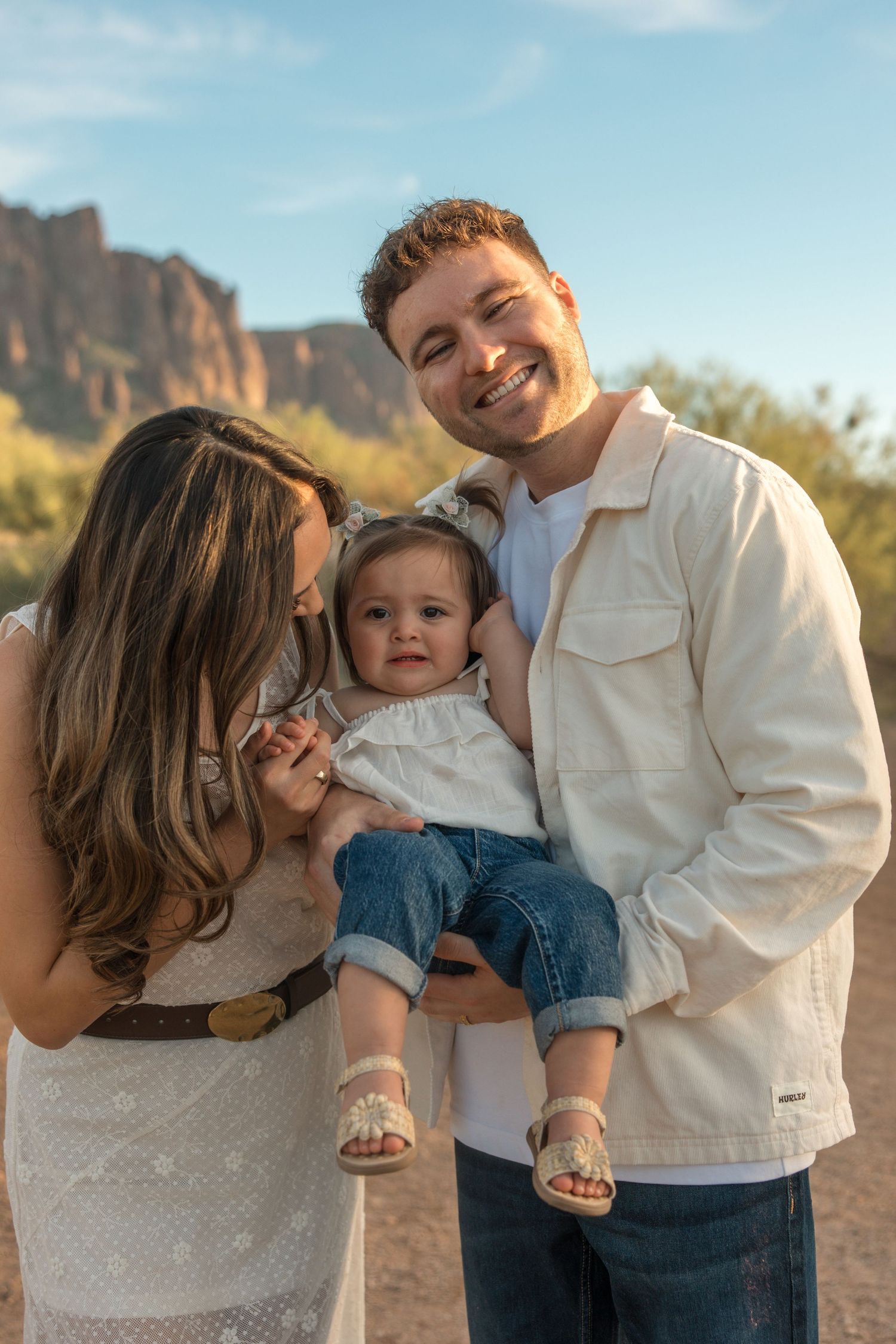 Happy family with dad holding baby girl as mom kisses her cheek in a desert landscape.