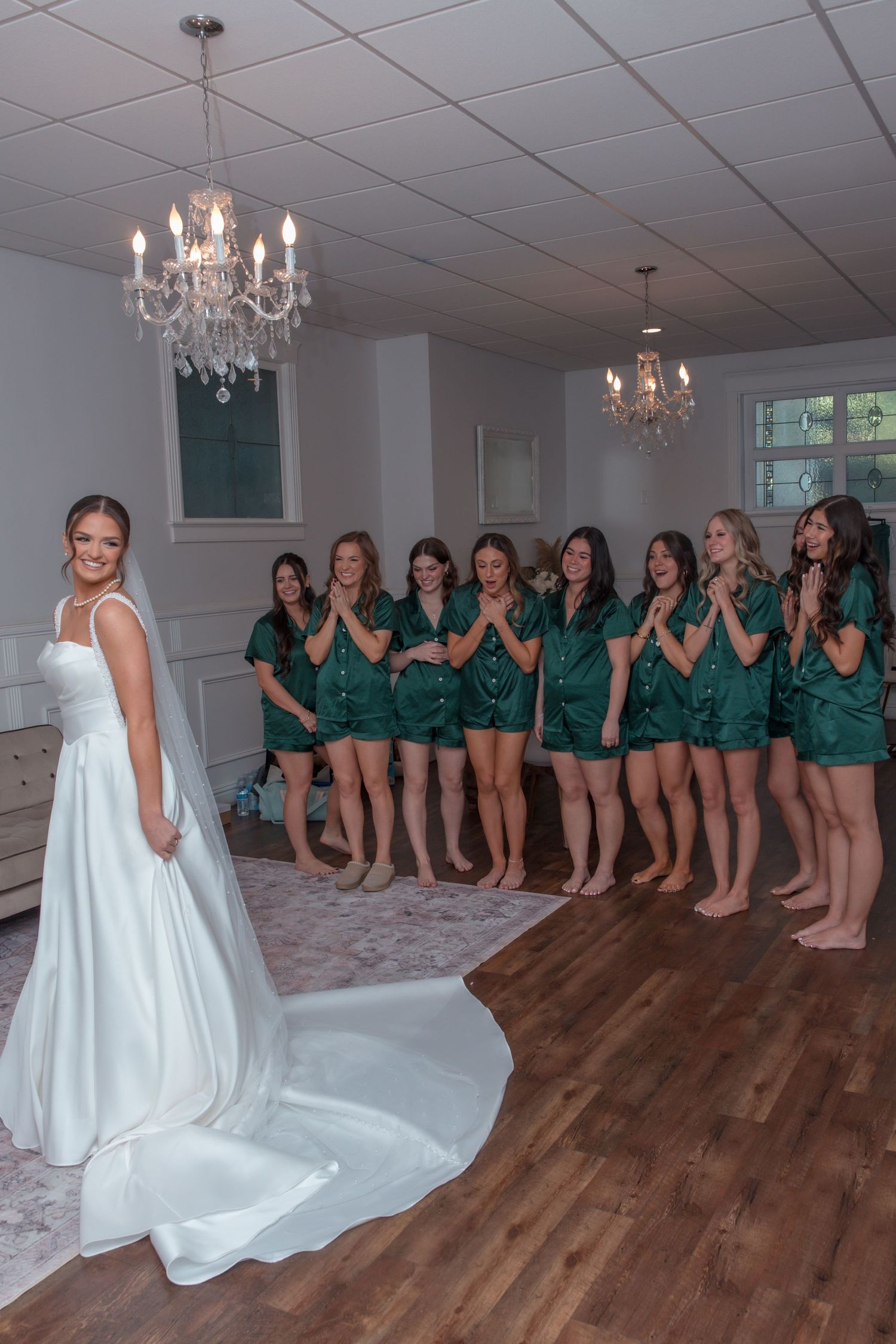 Bride in elegant white gown smiling at bridesmaids in matching green satin pajamas during first look.