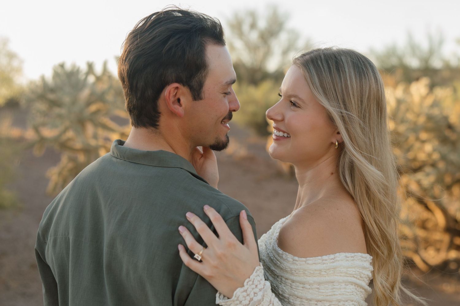 Couple smiling at each other during outdoor desert engagement photo session with cacti in background.