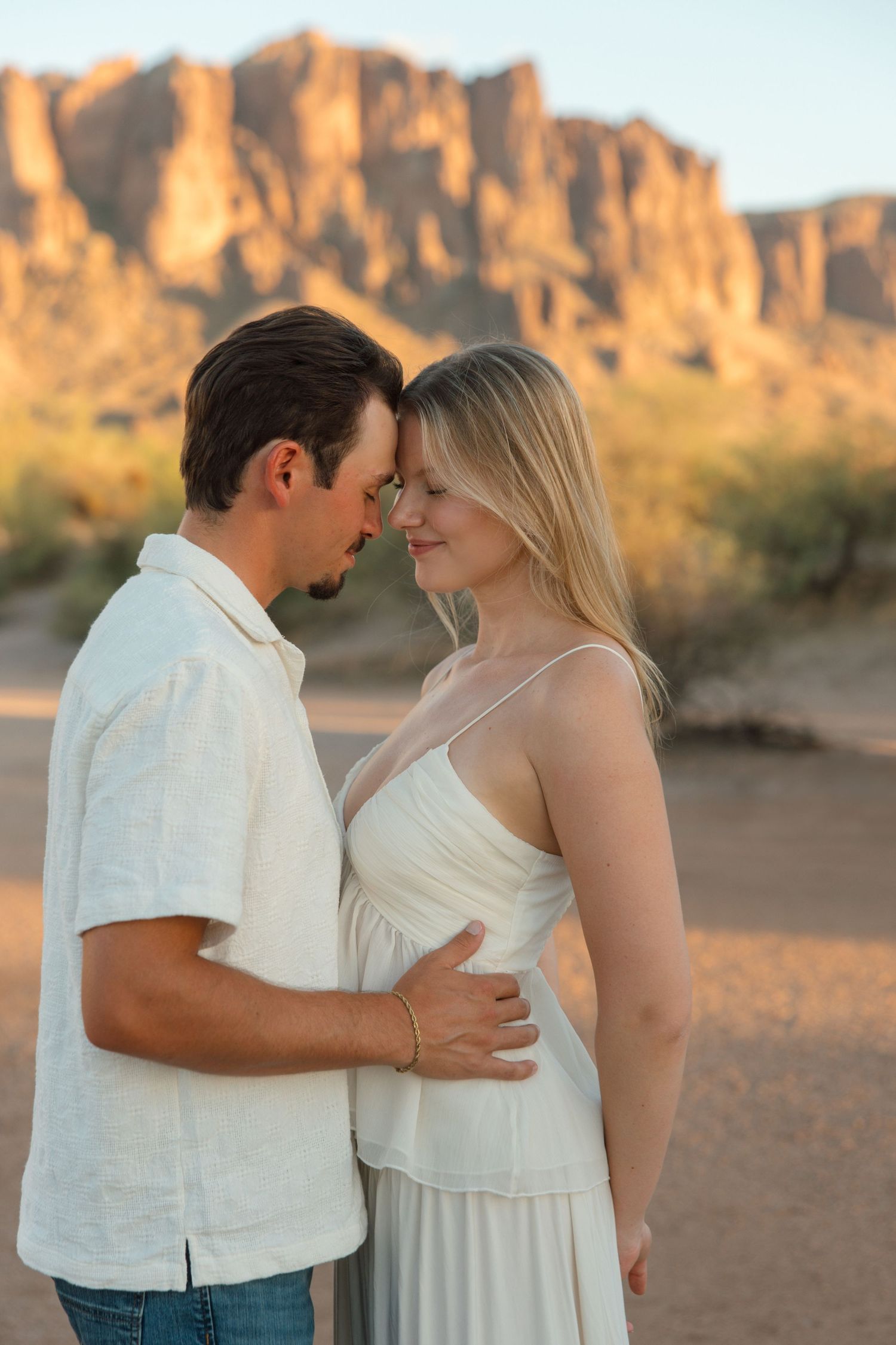 Couple in white attire sharing a romantic moment in the Arizona desert with Superstition Mountains at sunset.