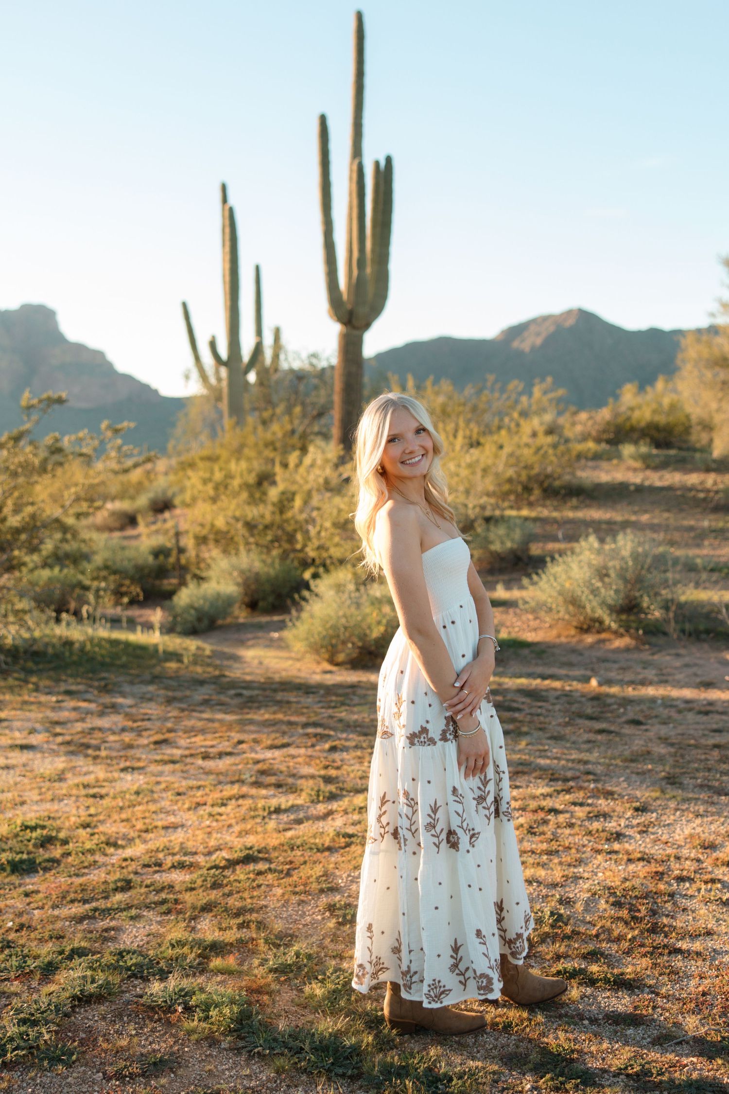 Smiling blonde woman in white floral maxi dress standing in Arizona desert with saguaro cactus and mountains at golden hour.