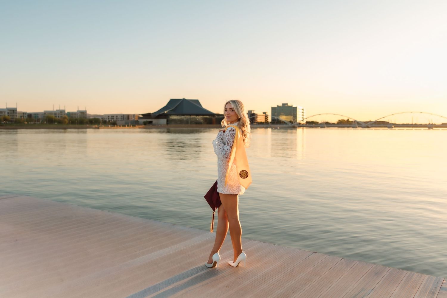 Woman in white outfit and hat walks on waterfront boardwalk at golden hour with city skyline behind her.