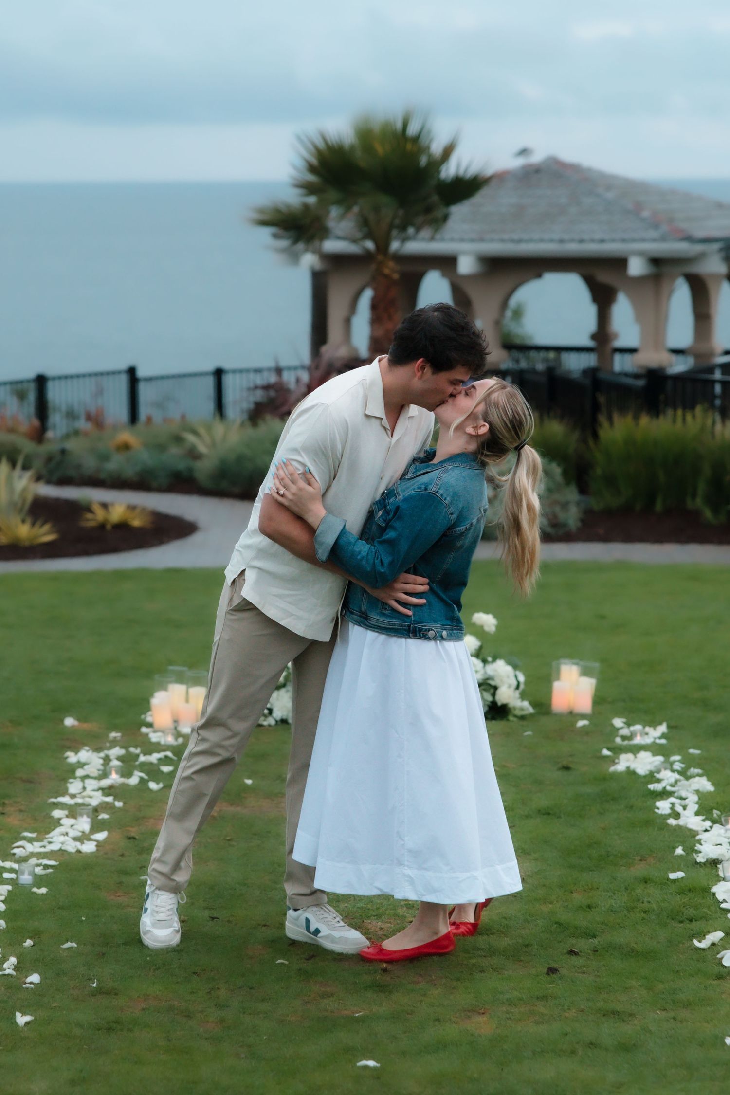 Newly engaged couple sharing passionate dip kiss on garden lawn with candles and white floral arrangements.
