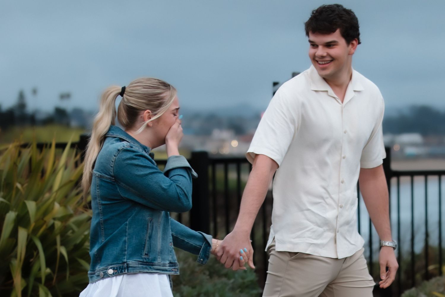 A couple holds hands outdoors, woman in denim jacket laughing shyly, man in white shirt smiling warmly at her.