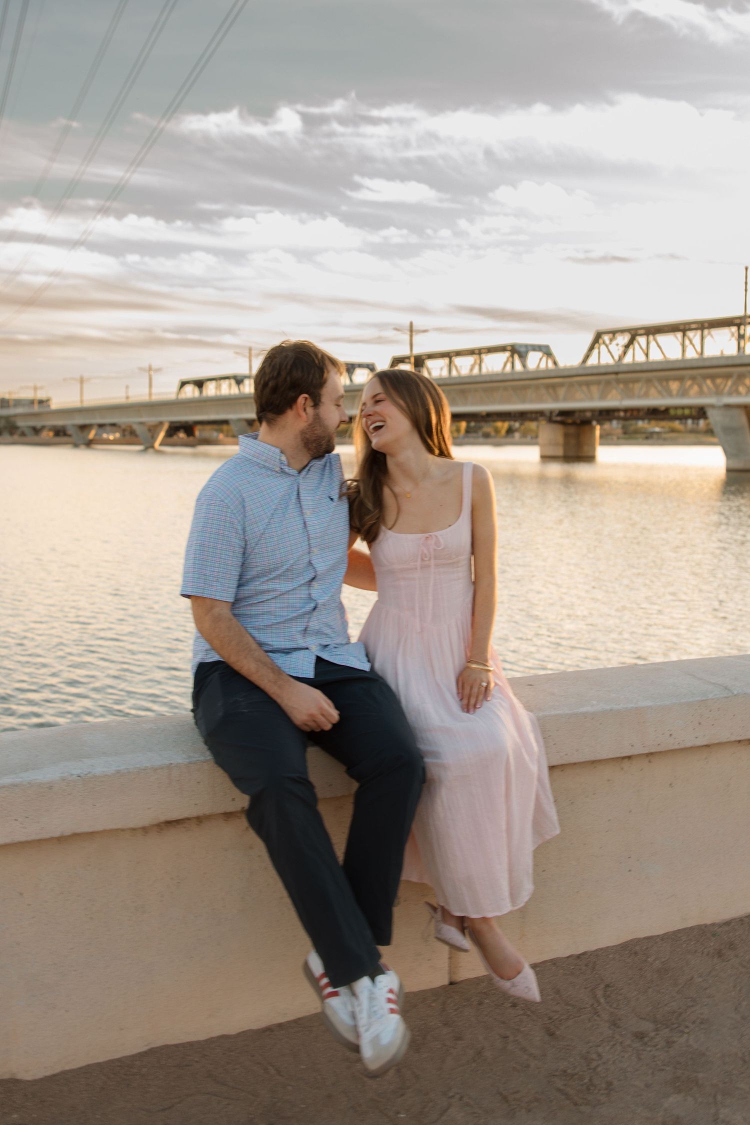 Engaged couple sitting together on waterfront wall laughing at sunset with bridge in background.