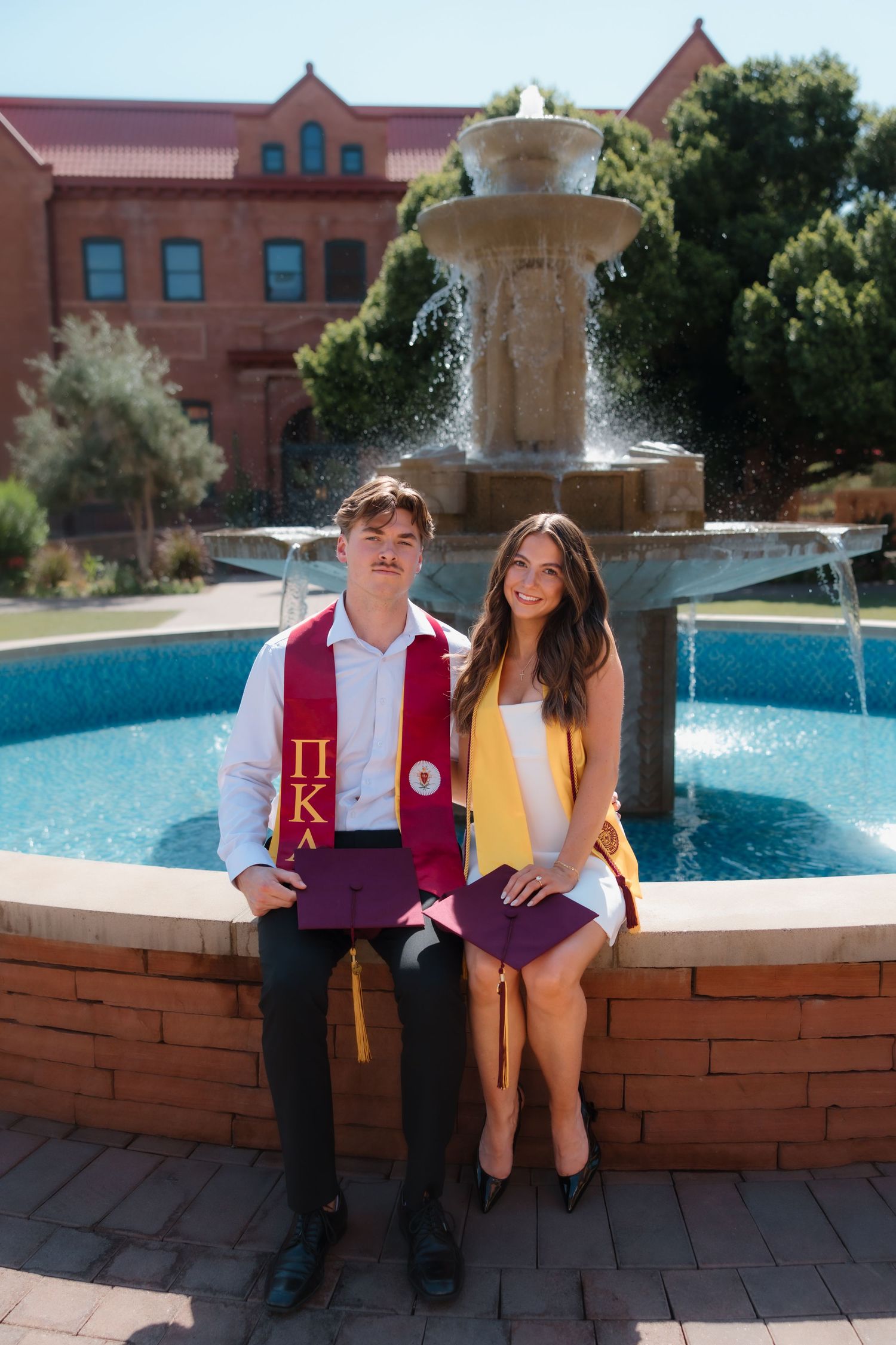 Two college graduates in honor stoles sit together smiling in front of a campus fountain.
