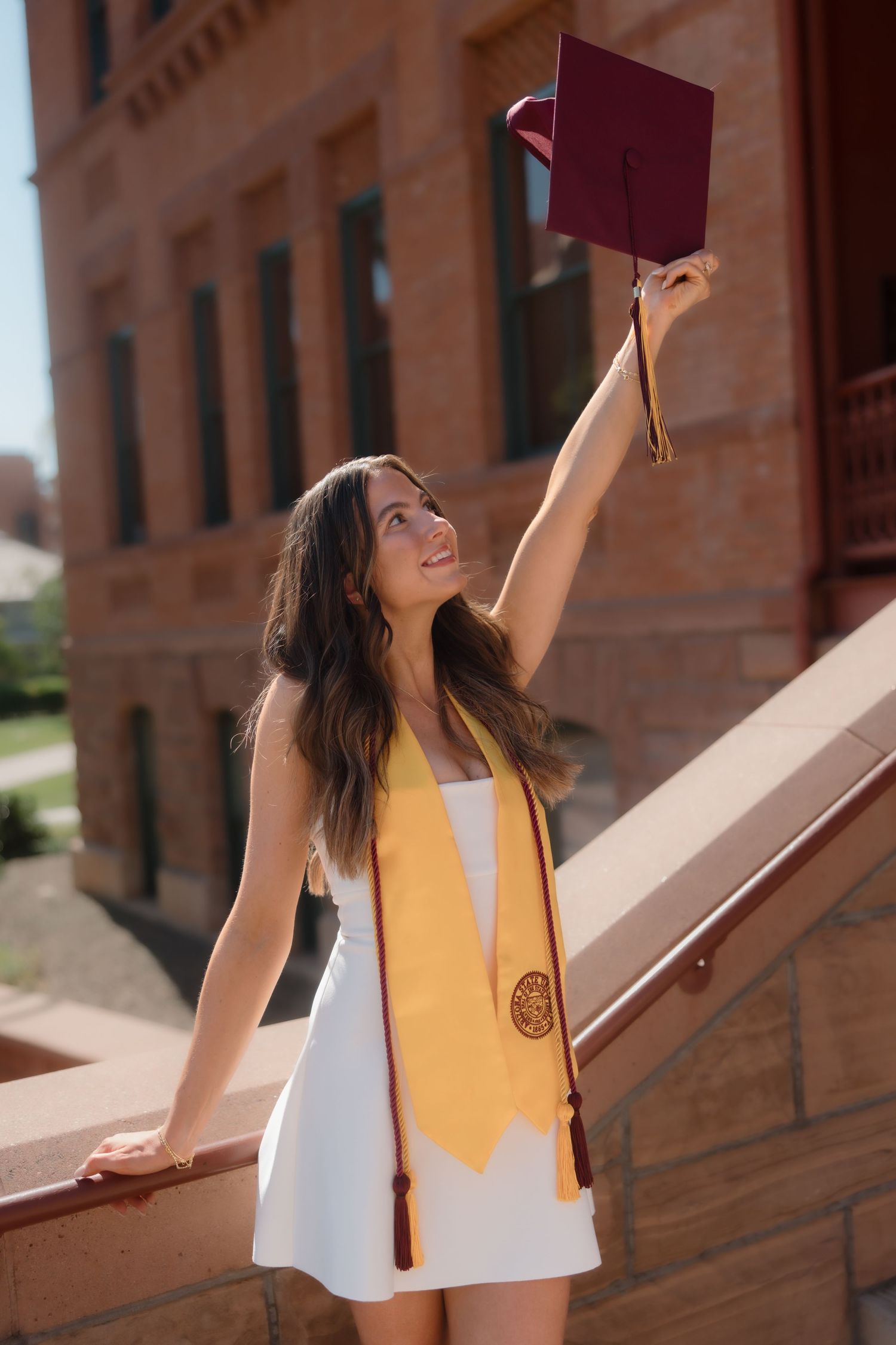 Smiling graduate in white dress raising maroon cap beside ASU brick building staircase.