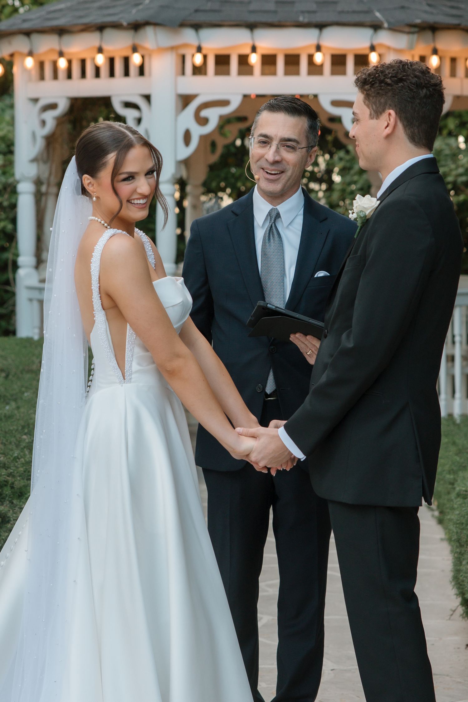 Smiling bride and groom hold hands at outdoor gazebo wedding ceremony with officiant laughing beside them.