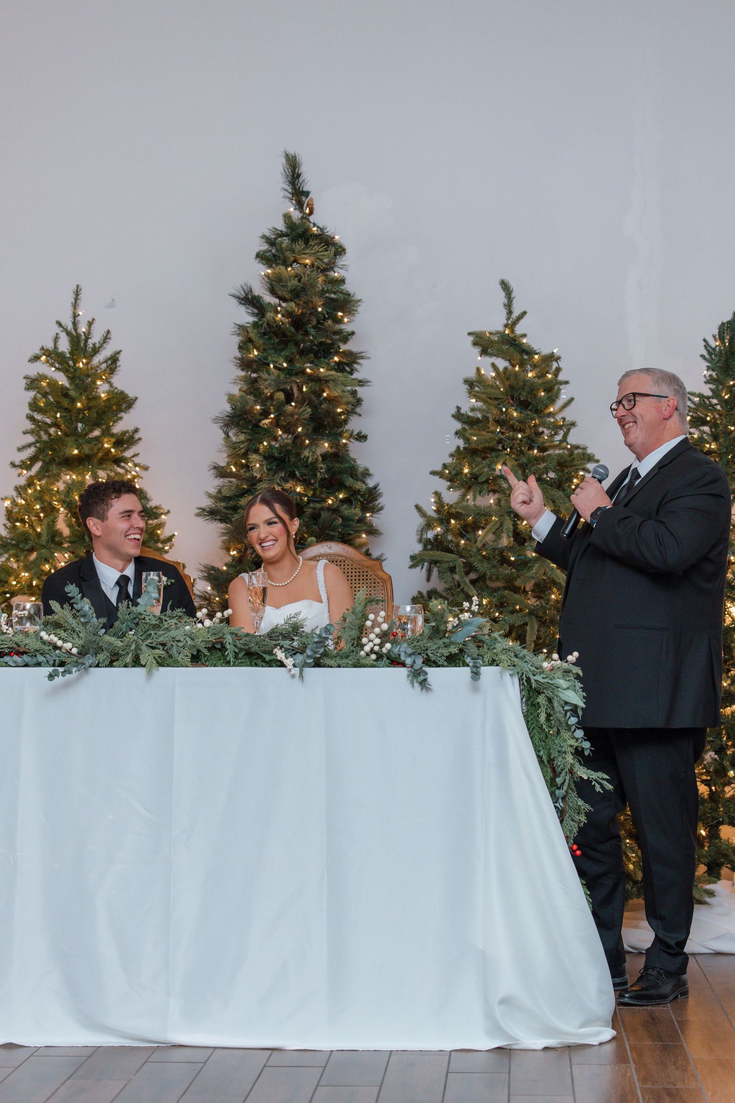 Older man giving wedding toast with microphone as bride and groom laugh at sweetheart table with Christmas trees.