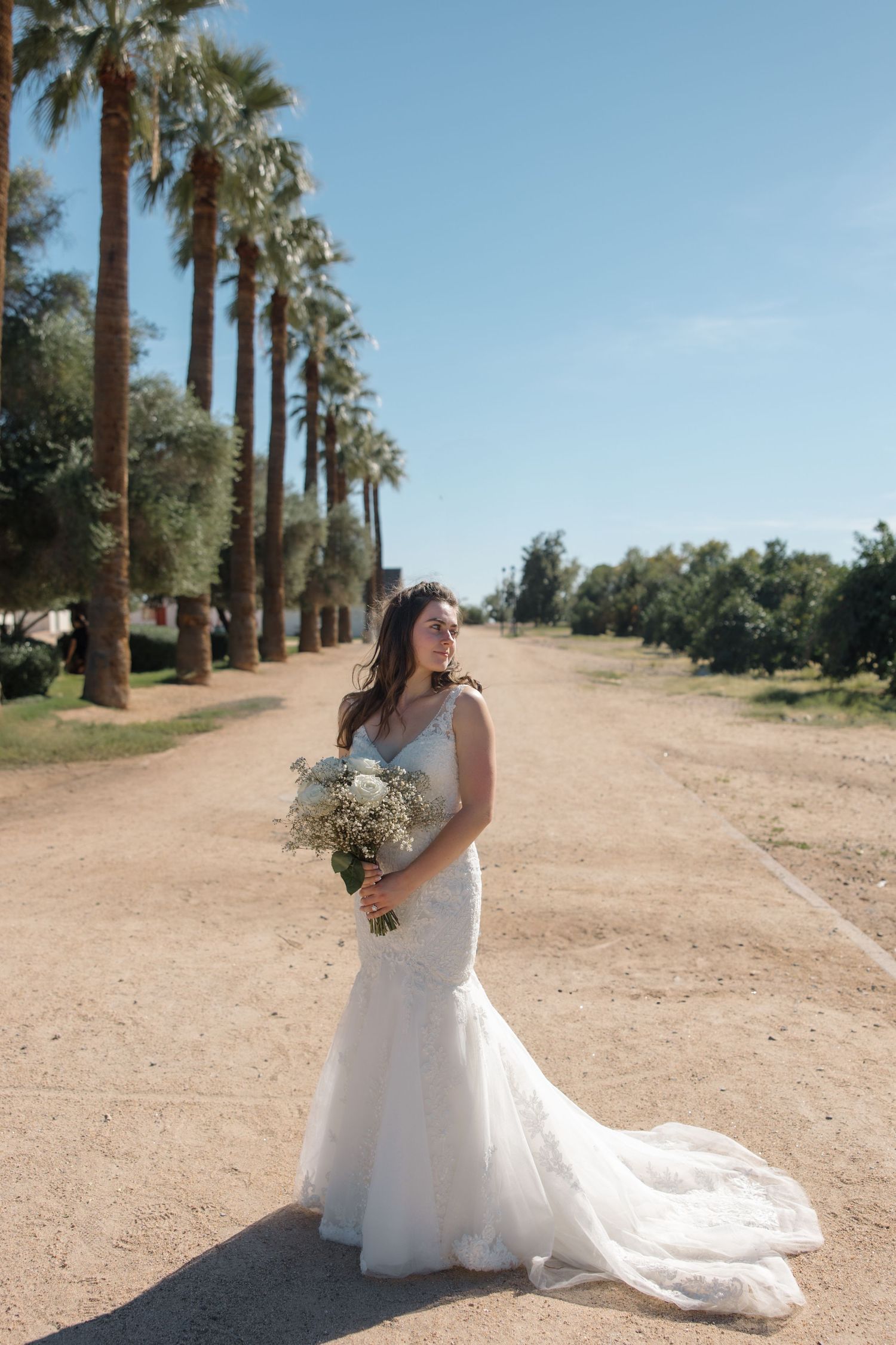 Elegant bride in mermaid lace gown holding white bouquet standing on sandy path lined with tall palm trees.