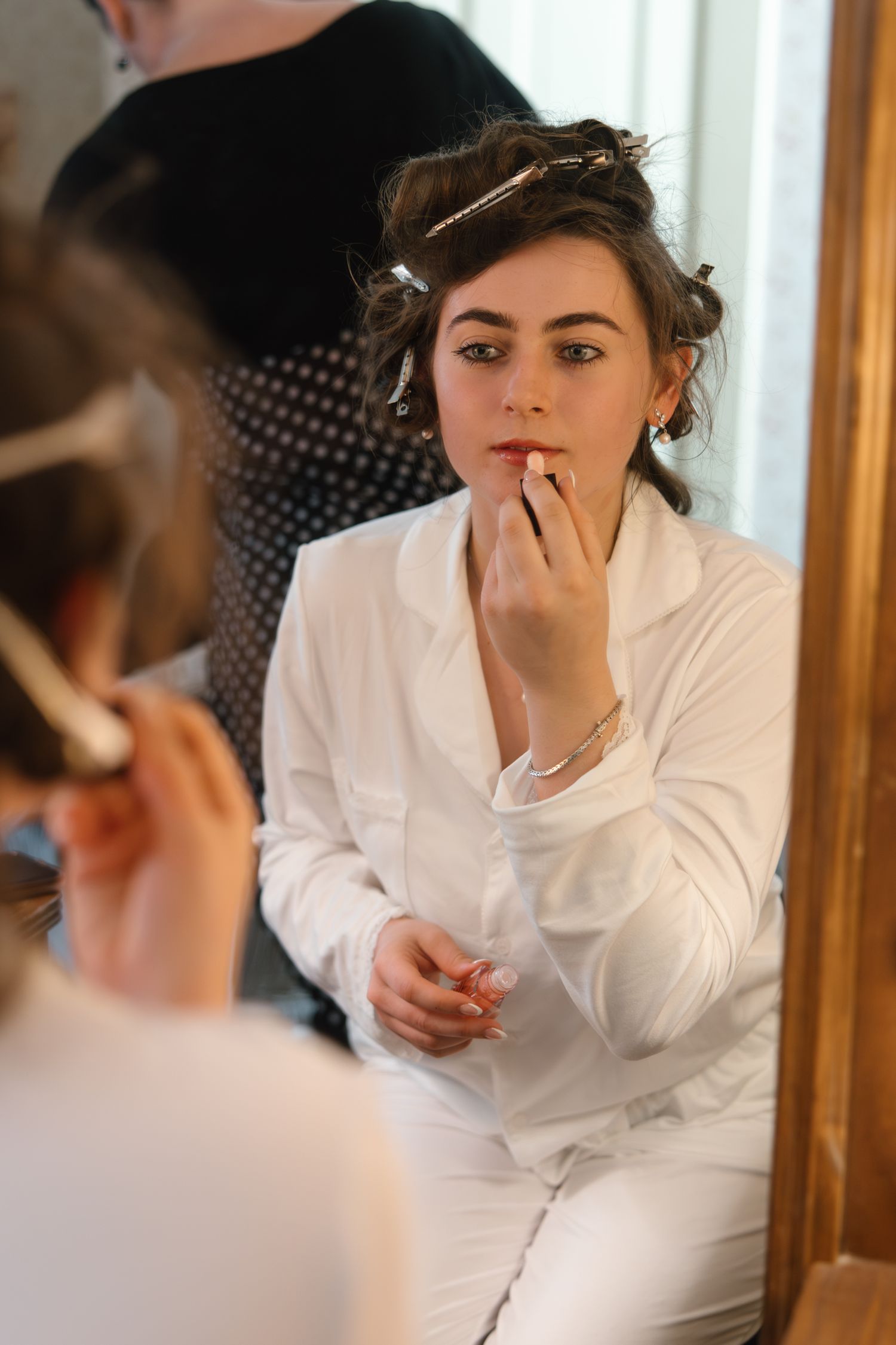 Bride applying lipstick while sitting before mirror with hair in rollers during wedding preparations.