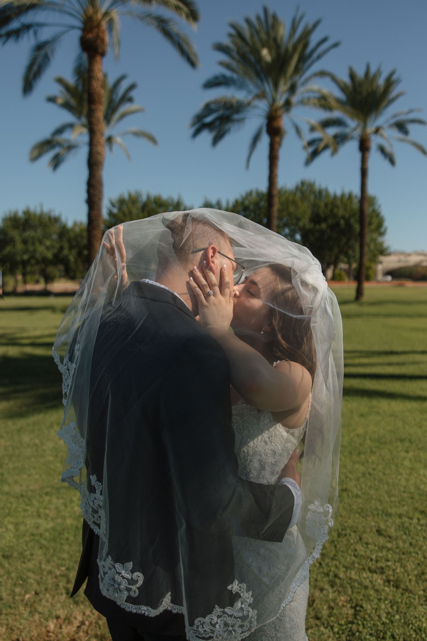 Bride and groom share a romantic kiss under a lace wedding veil on a green lawn with palm trees.