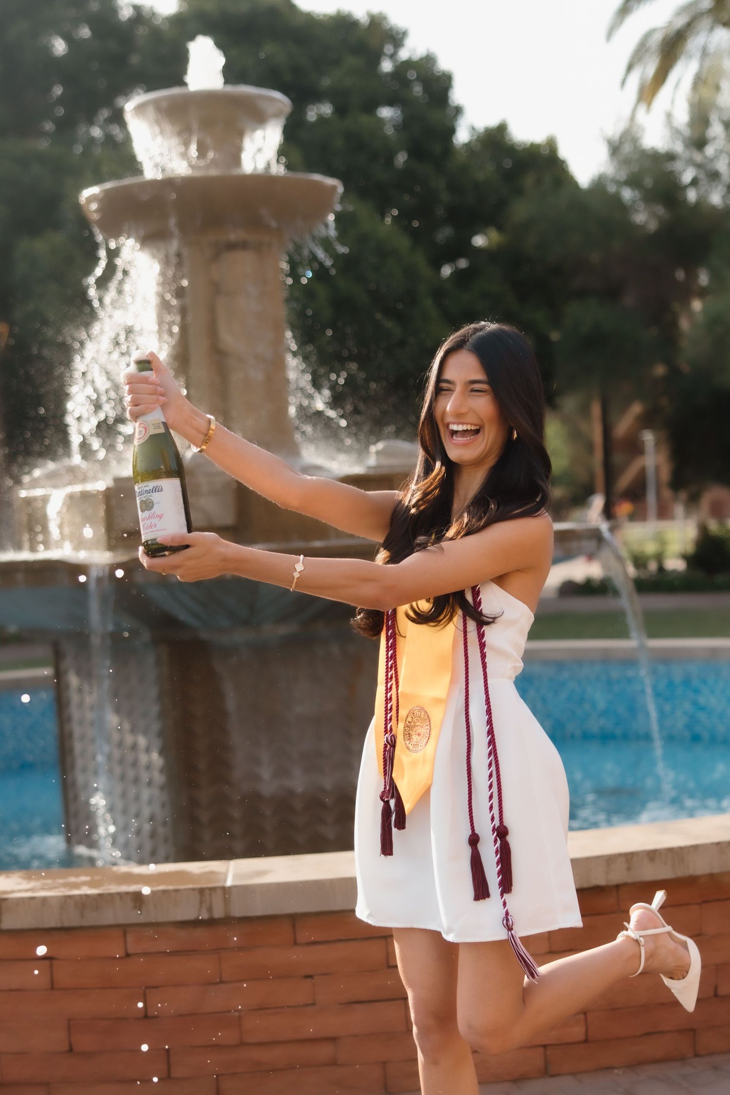 Smiling ASU graduate in white dress popping champagne bottle by campus fountain wearing maroon and gold graduation sash.
