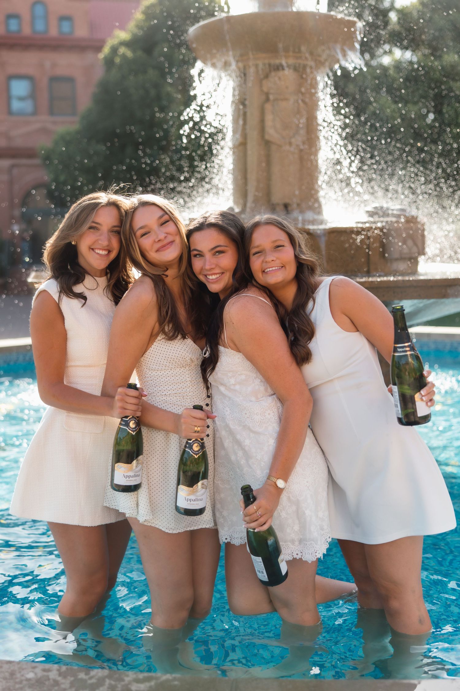 Four smiling women in white dresses hold champagne bottles while standing in a blue fountain.