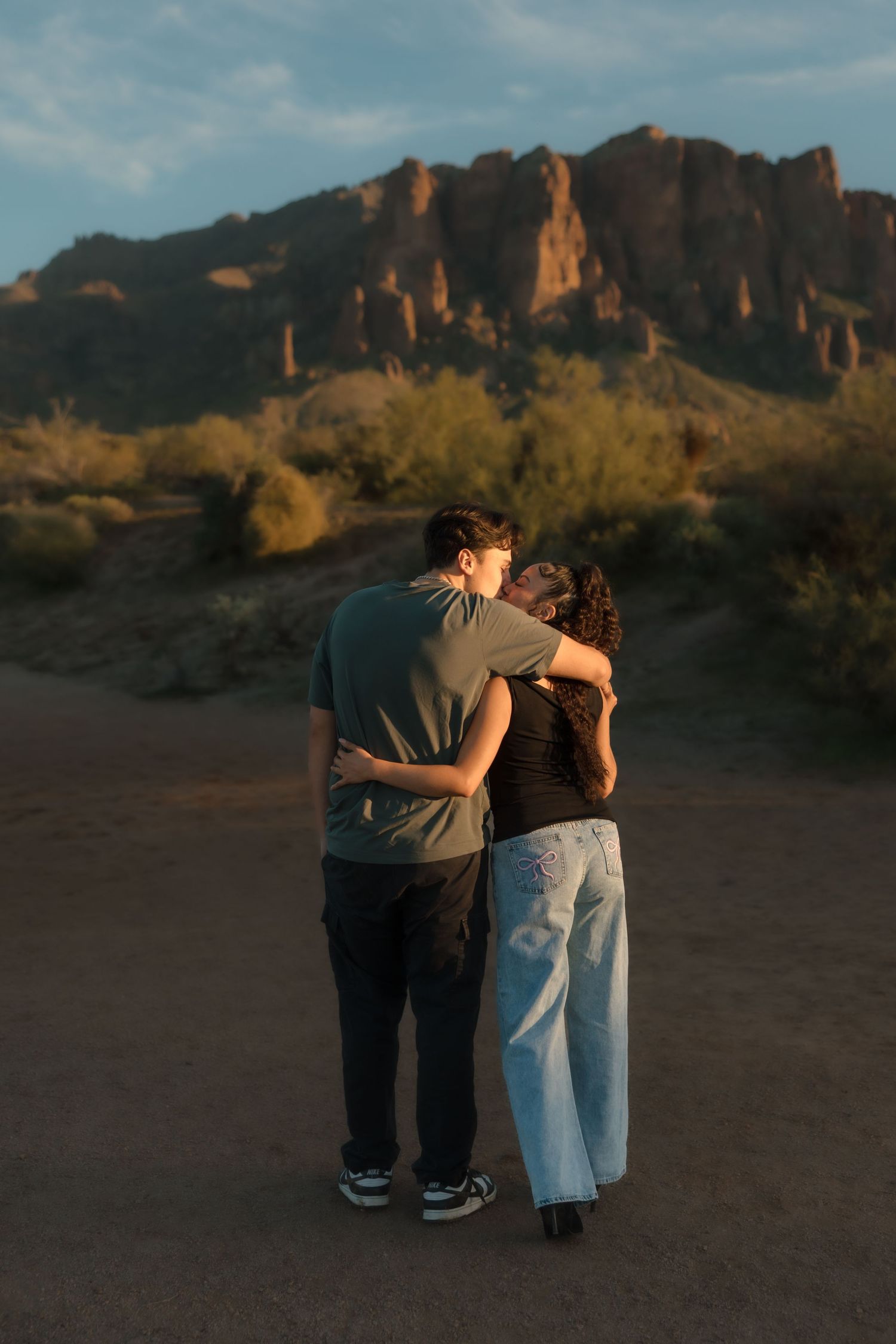 A couple shares a romantic kiss in a desert landscape with towering rocky mountains at sunset.
