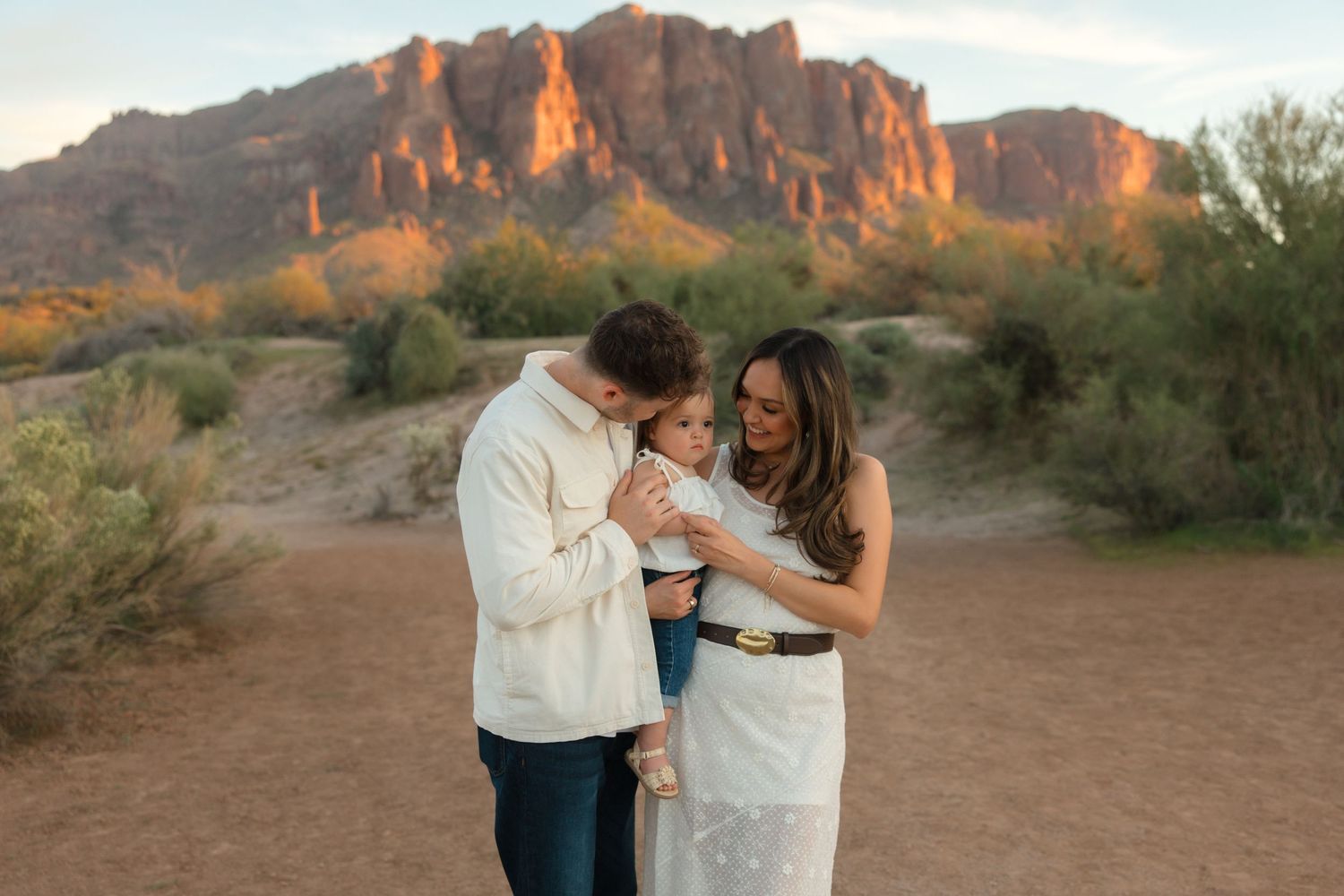 Family of three sharing a sweet moment together in Arizona desert with glowing mountain backdrop.