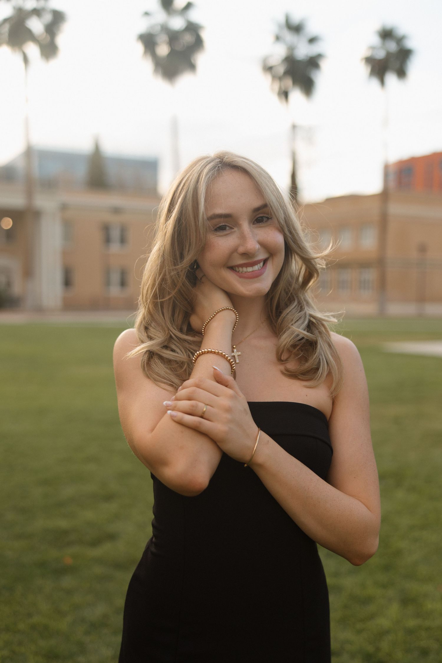 Smiling blonde woman in black strapless dress poses outdoors on green lawn with palm trees and building in background.