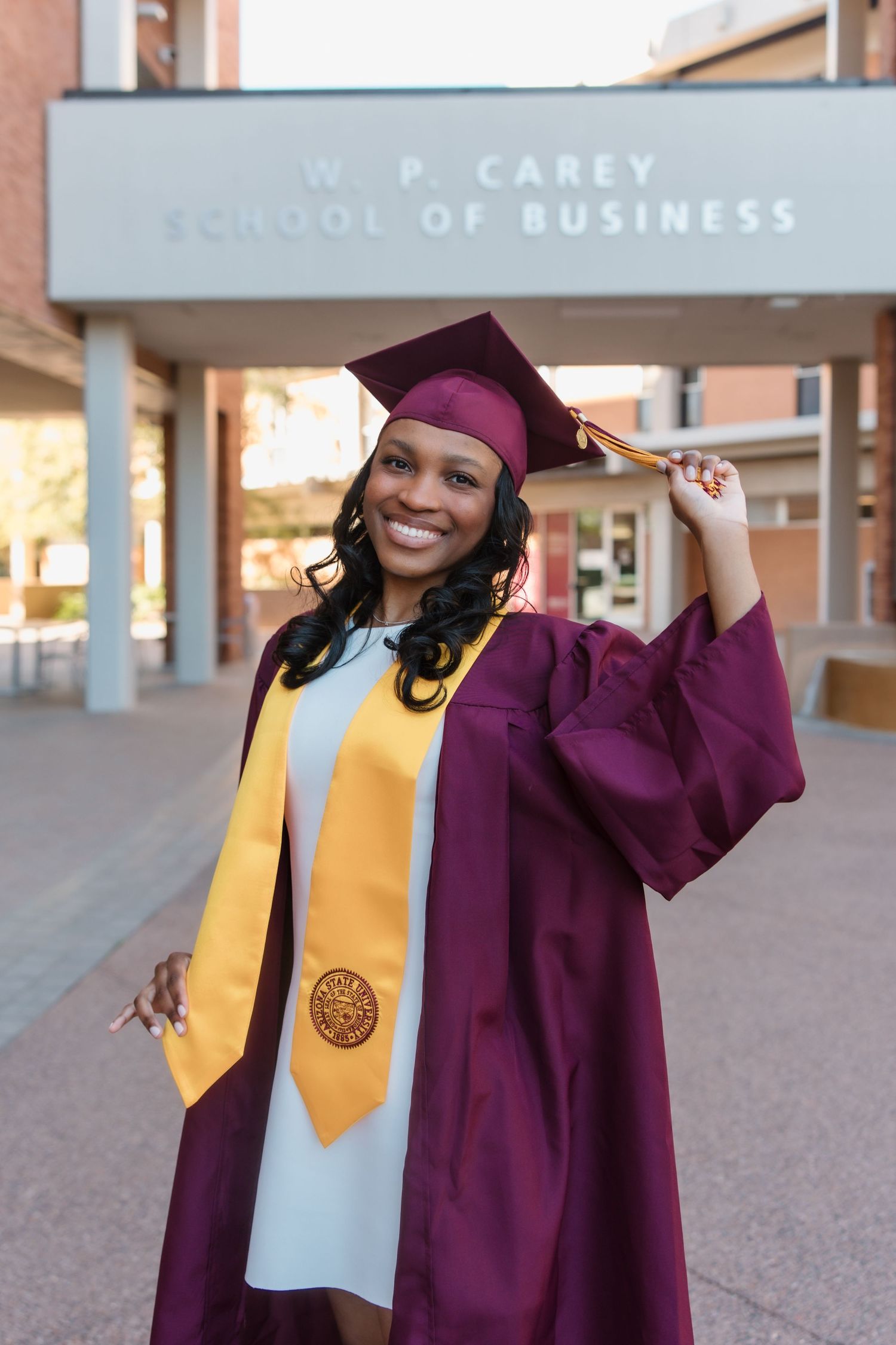 A smiling graduate in maroon cap and gown poses in front of the W.P. Carey School of Business building.